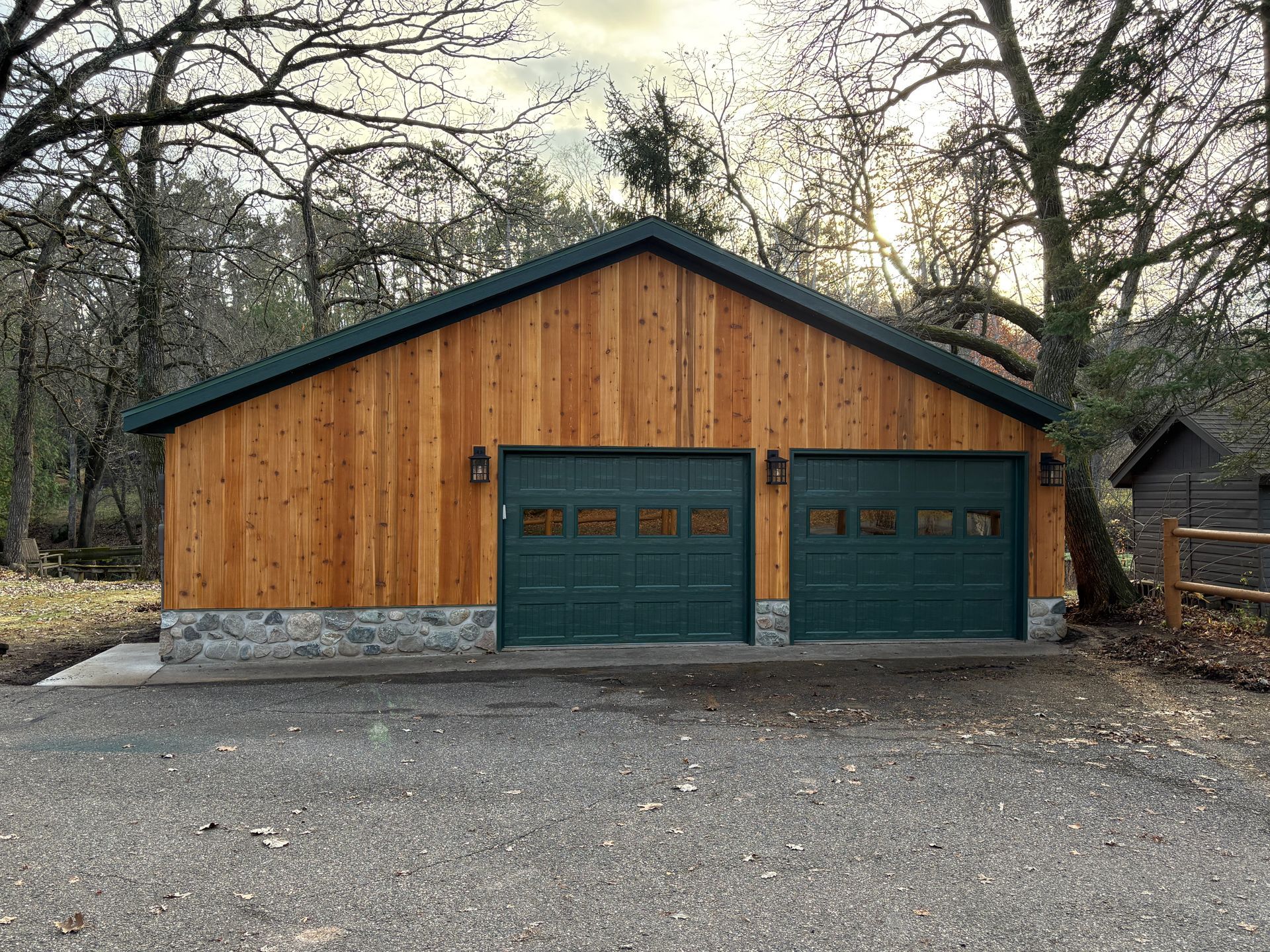 Two-car garage with green doors, cedar siding, and a dark green roof, set on a gravel driveway.