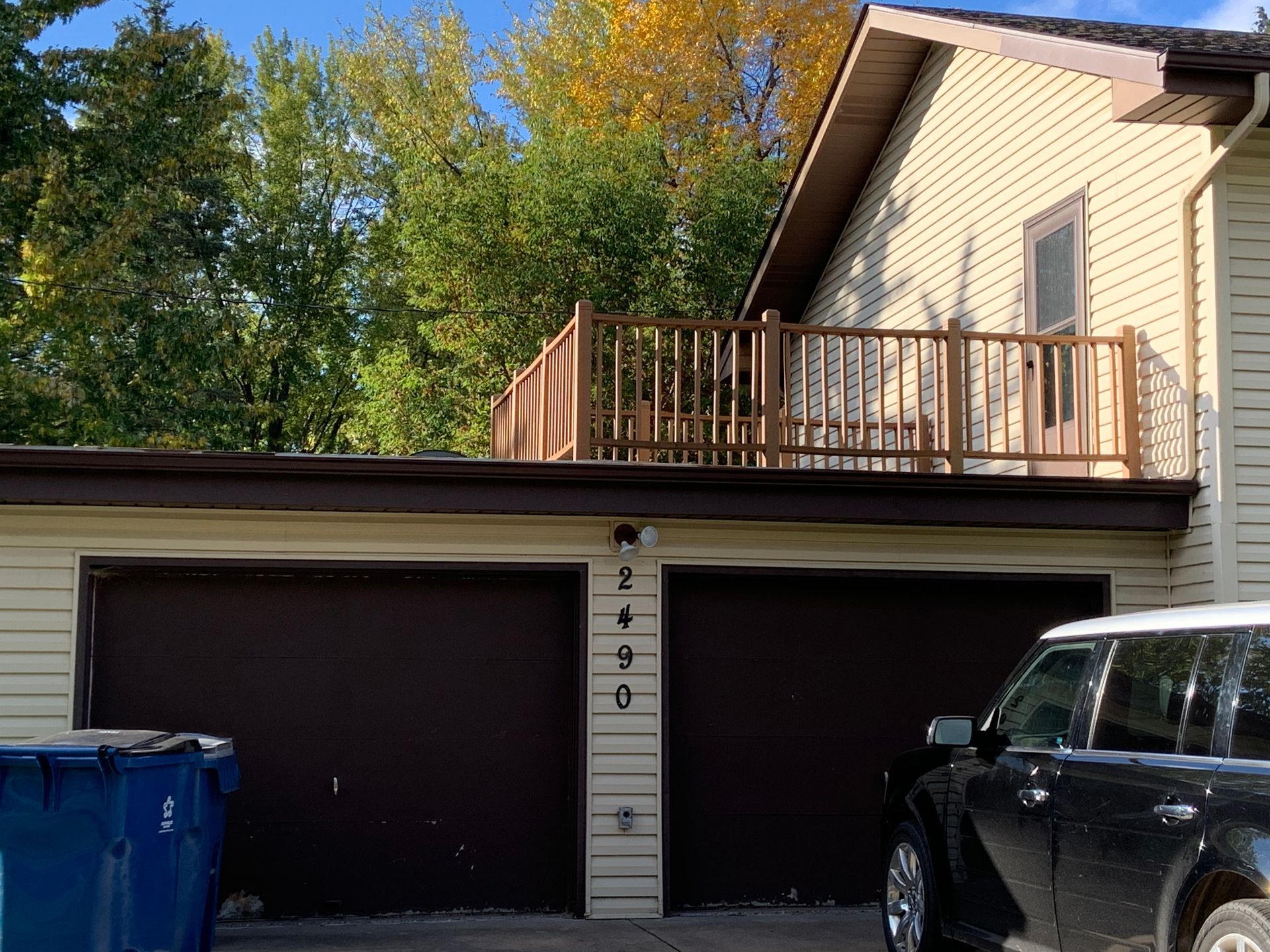 A two-car garage with brown doors, tan siding, the address 2490, and a wooden balcony above, parked car, and blue bin.