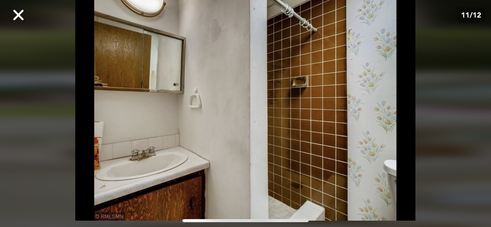 Small bathroom with a sink, mirror cabinet, and brown-tiled shower next to floral wallpaper.