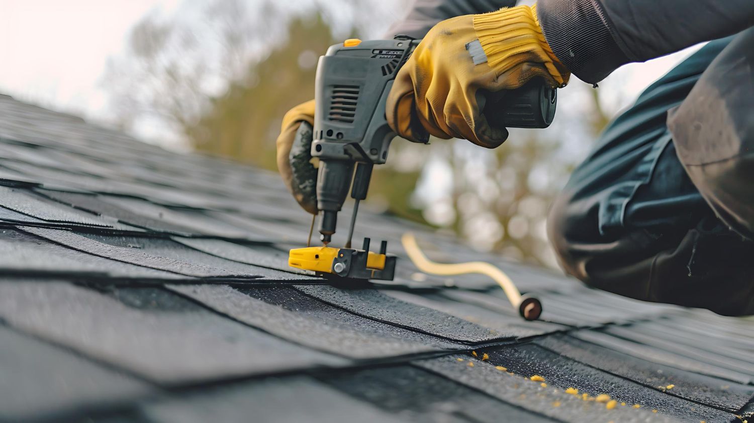 Worker fastening shingles on a roof with a nail gun, wearing yellow gloves