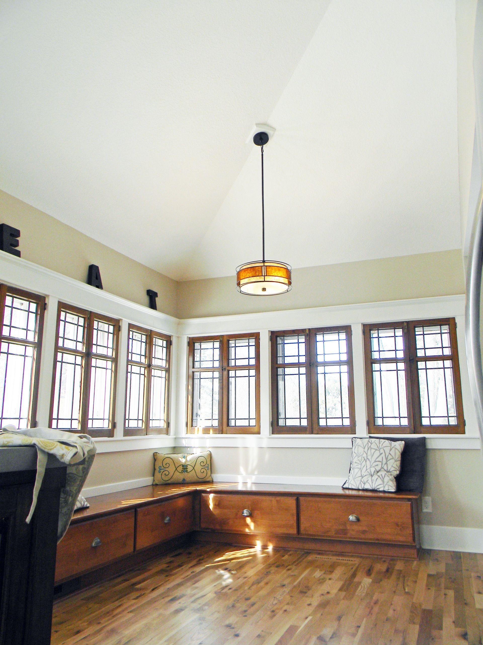 Sunlit built-in window seat in a room with hardwood floors, accented by wooden window frames, light fixture, and beige walls.