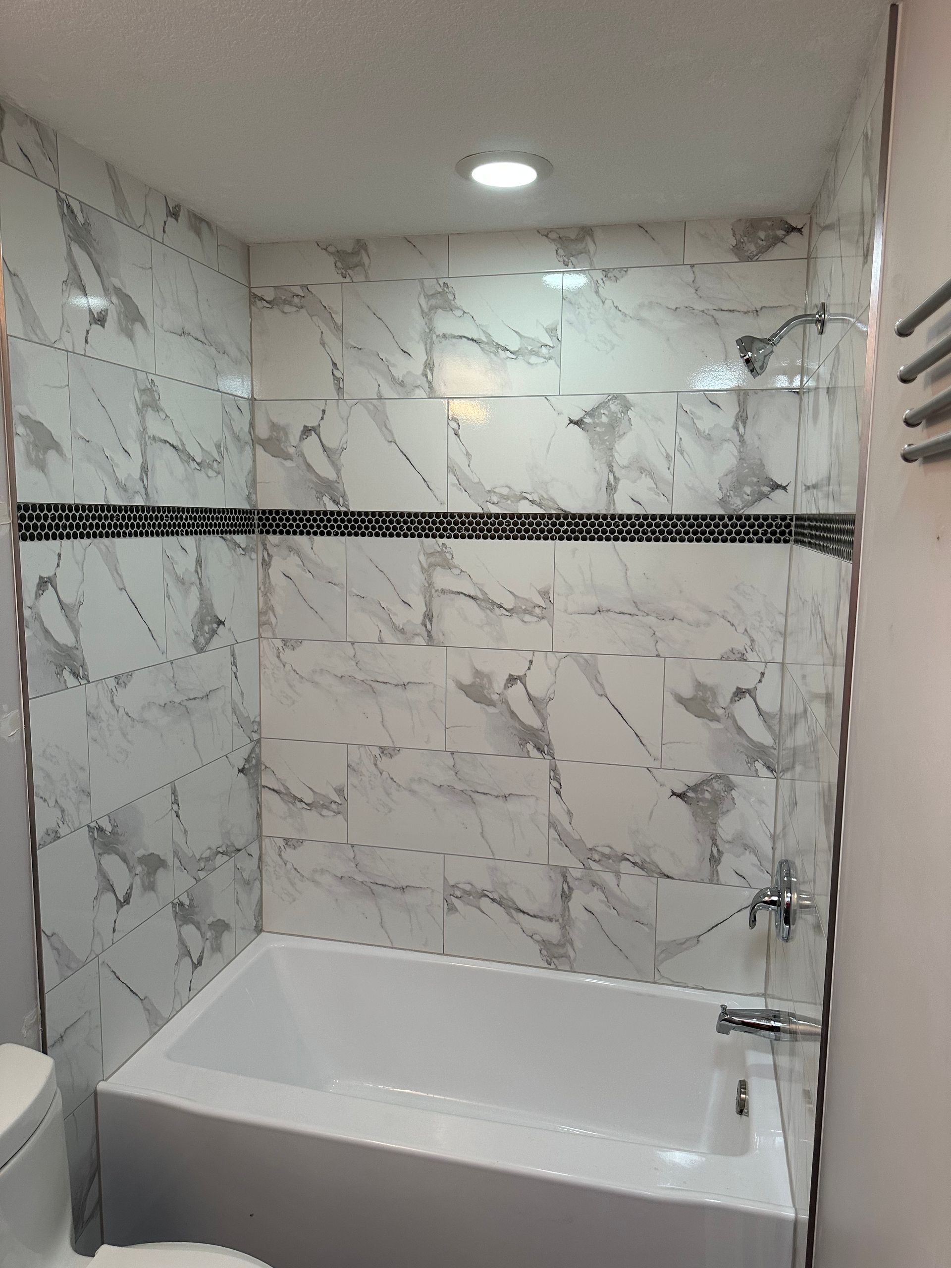 Tiled shower with white tub, marble-like walls, black accent tile, and overhead lighting.