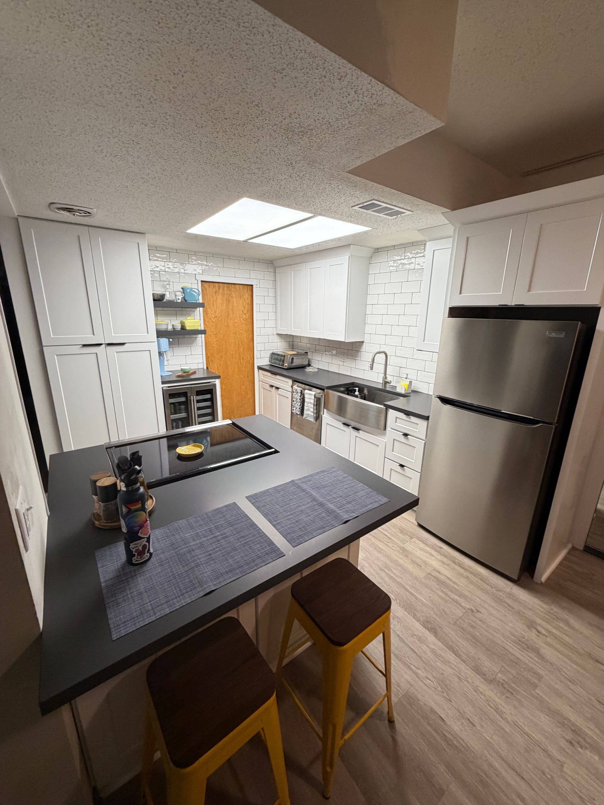 Modern kitchen with white cabinets, island, stainless steel refrigerator, and skylight. Two yellow stools at the island.