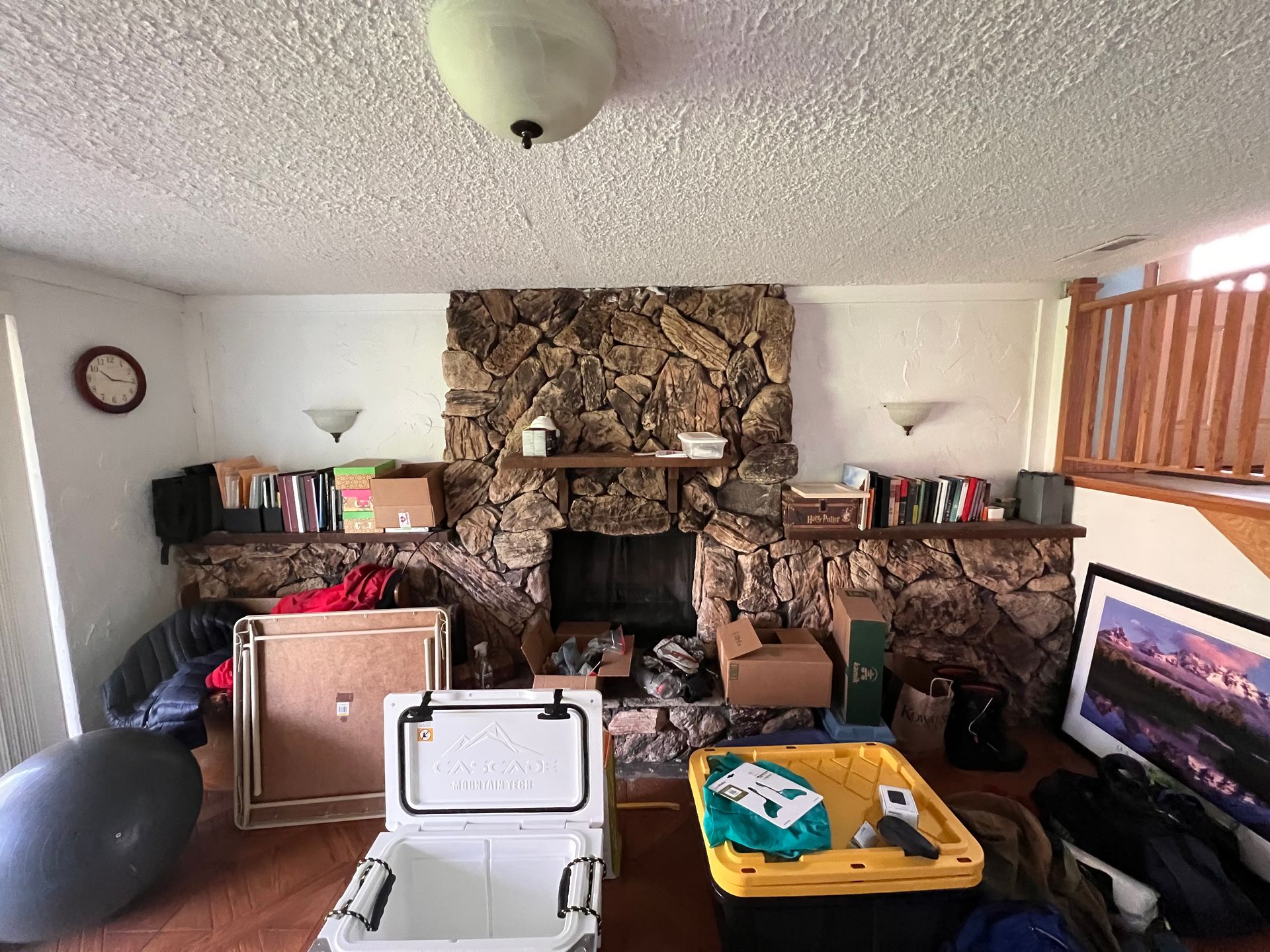 A cluttered living room with a stone fireplace, boxes, and a cooler.