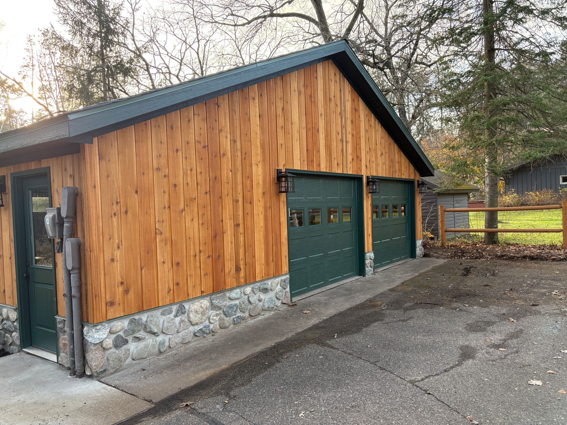 Cedar-sided garage with two green garage doors, green roof, and stone foundation. Driveway slopes up.
