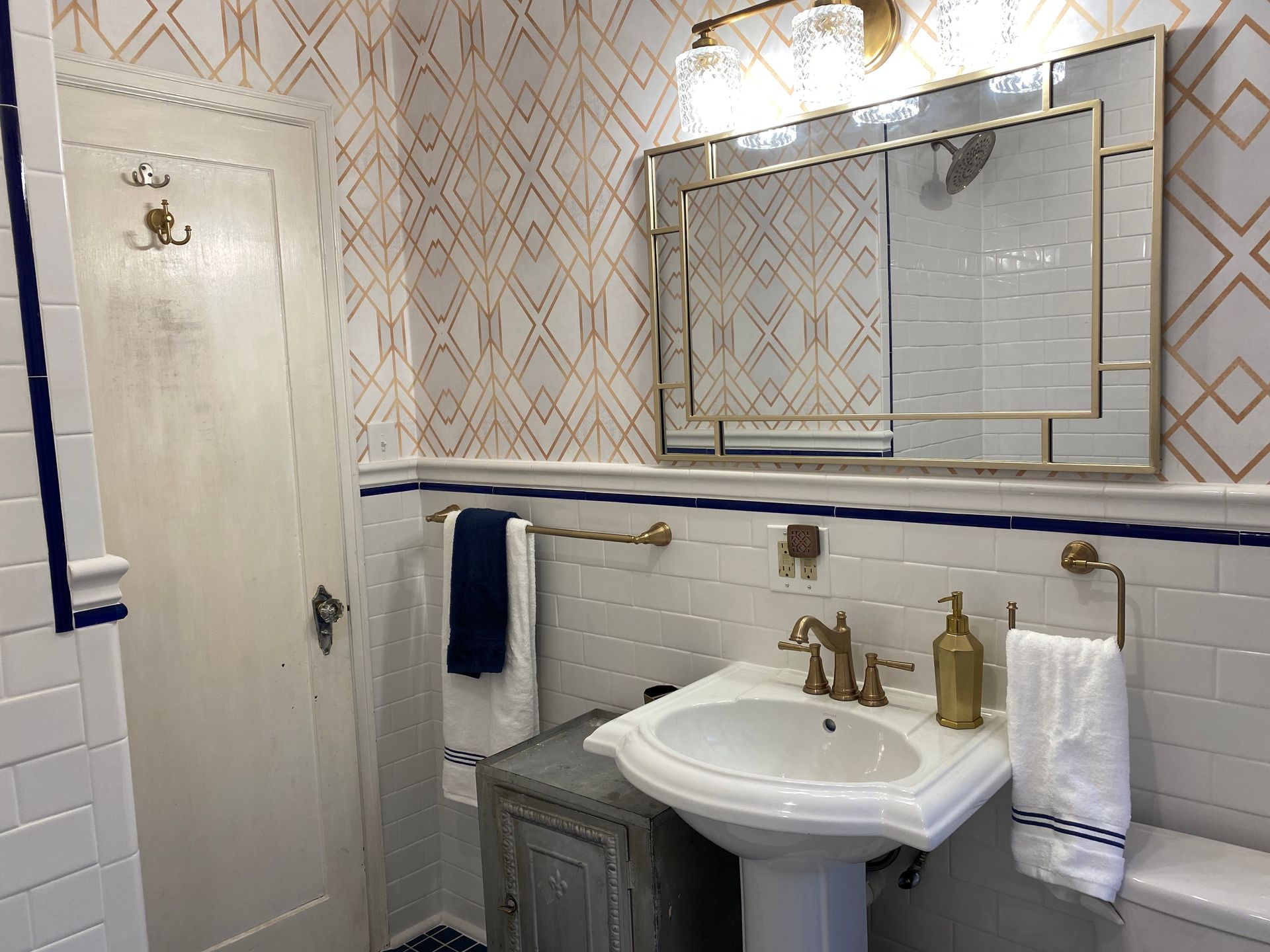 Bathroom with white and gold patterned wallpaper, pedestal sink, gold fixtures, and a navy blue accent.