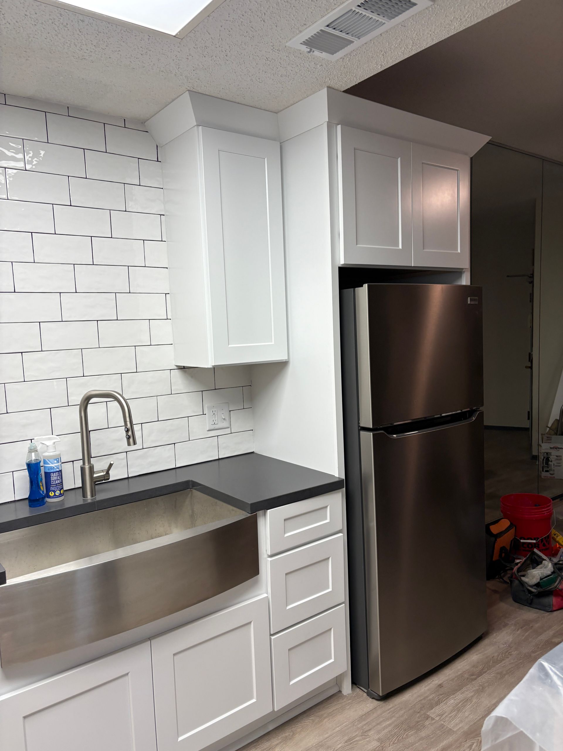 White kitchen cabinets and sink, stainless steel refrigerator and faucet, dark countertop, and decorative tile backsplash.