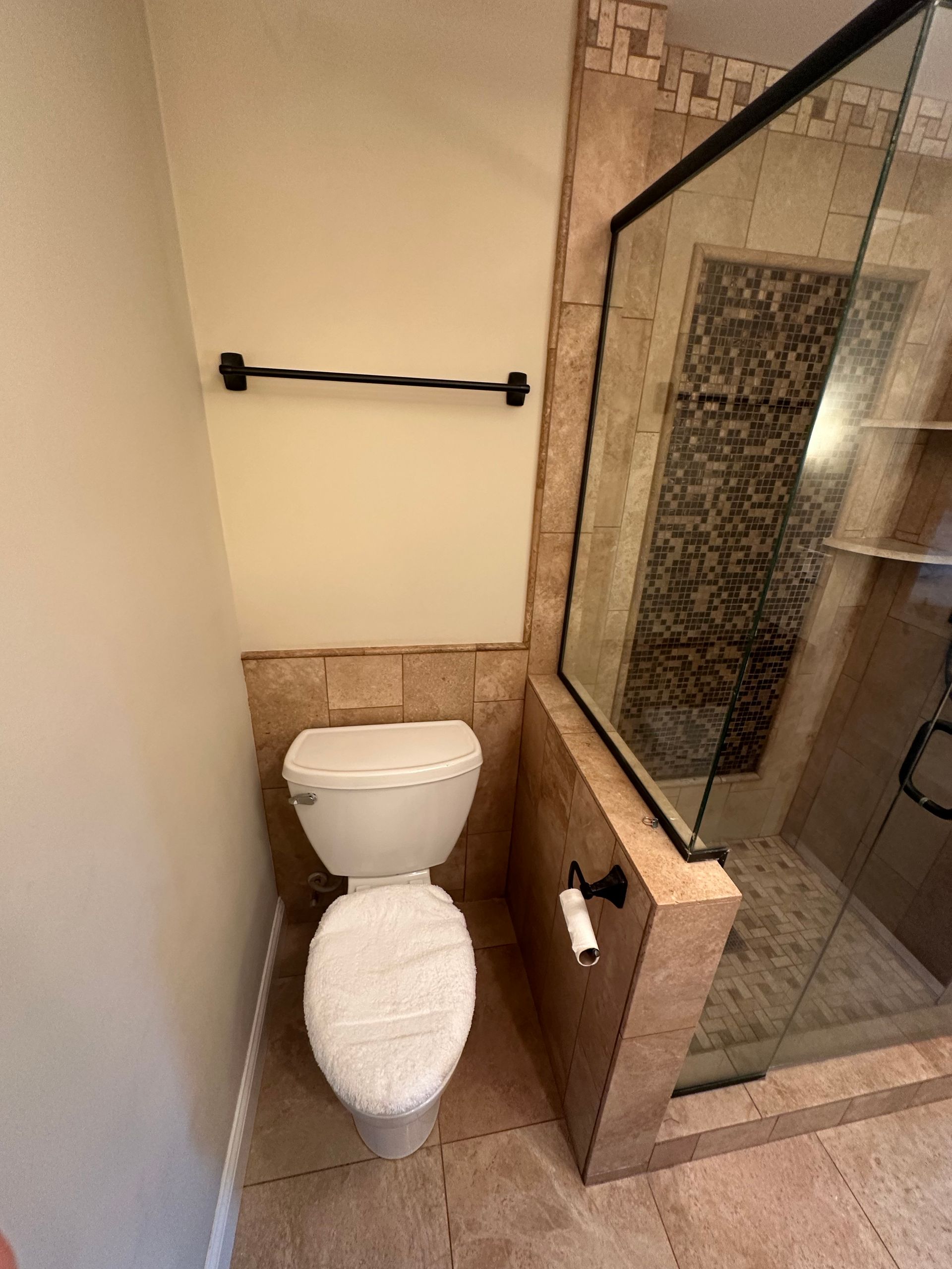 Toilet next to a glass shower in a bathroom with tan tile and a black towel rack.