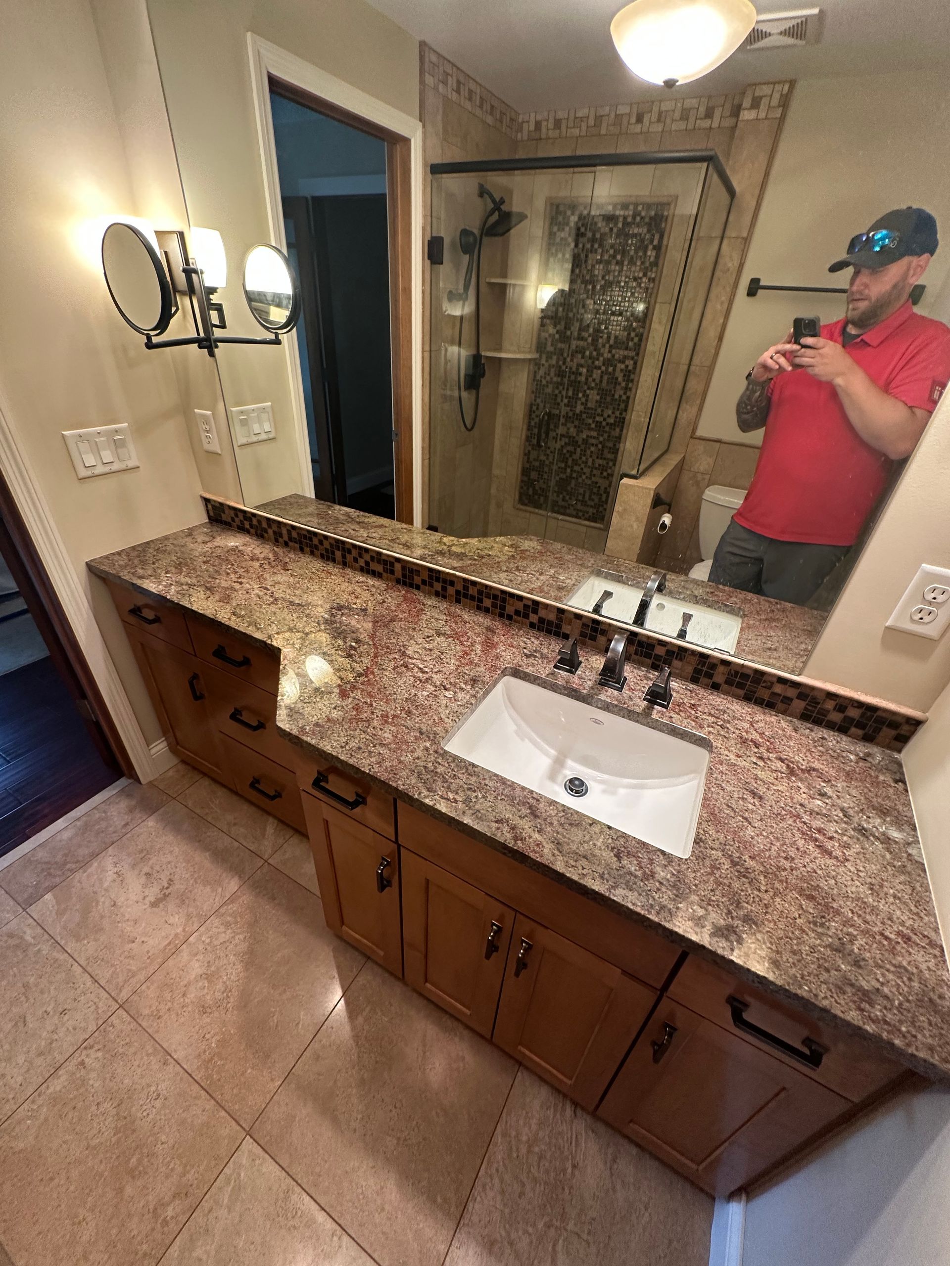 Bathroom with brown cabinets, granite countertop, white sink, and a man taking a photo in the mirror.