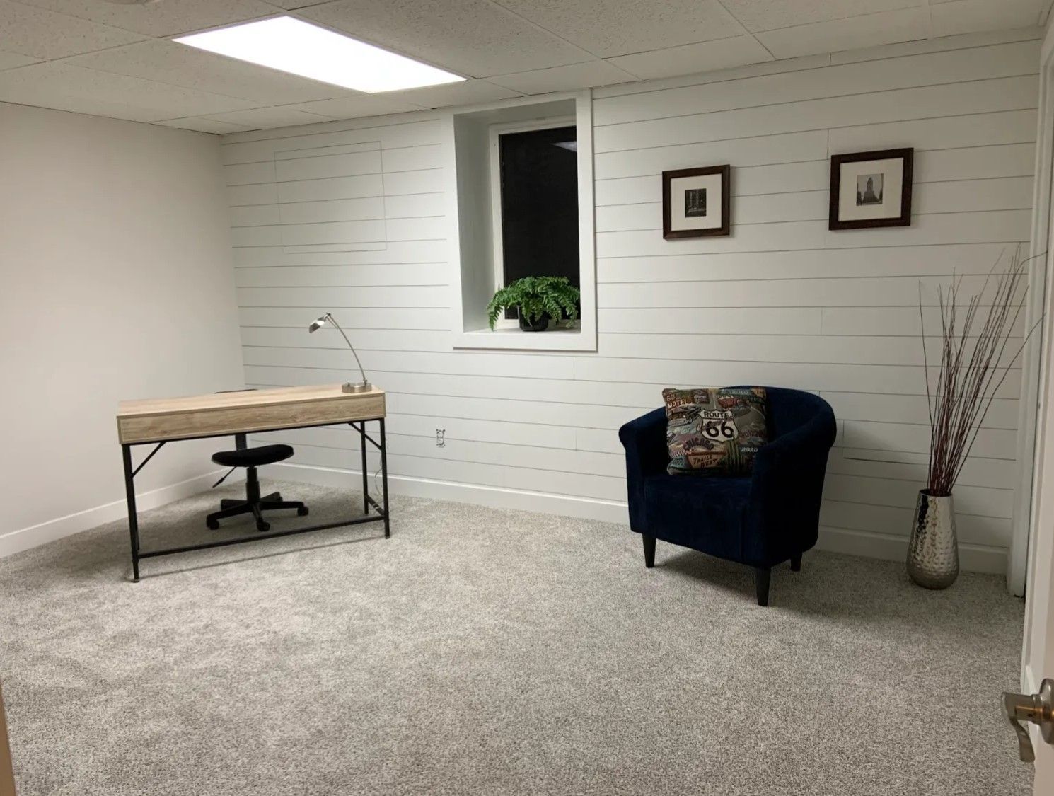 Office with desk, chair, blue armchair, window, and decorative items. White walls and gray carpet.