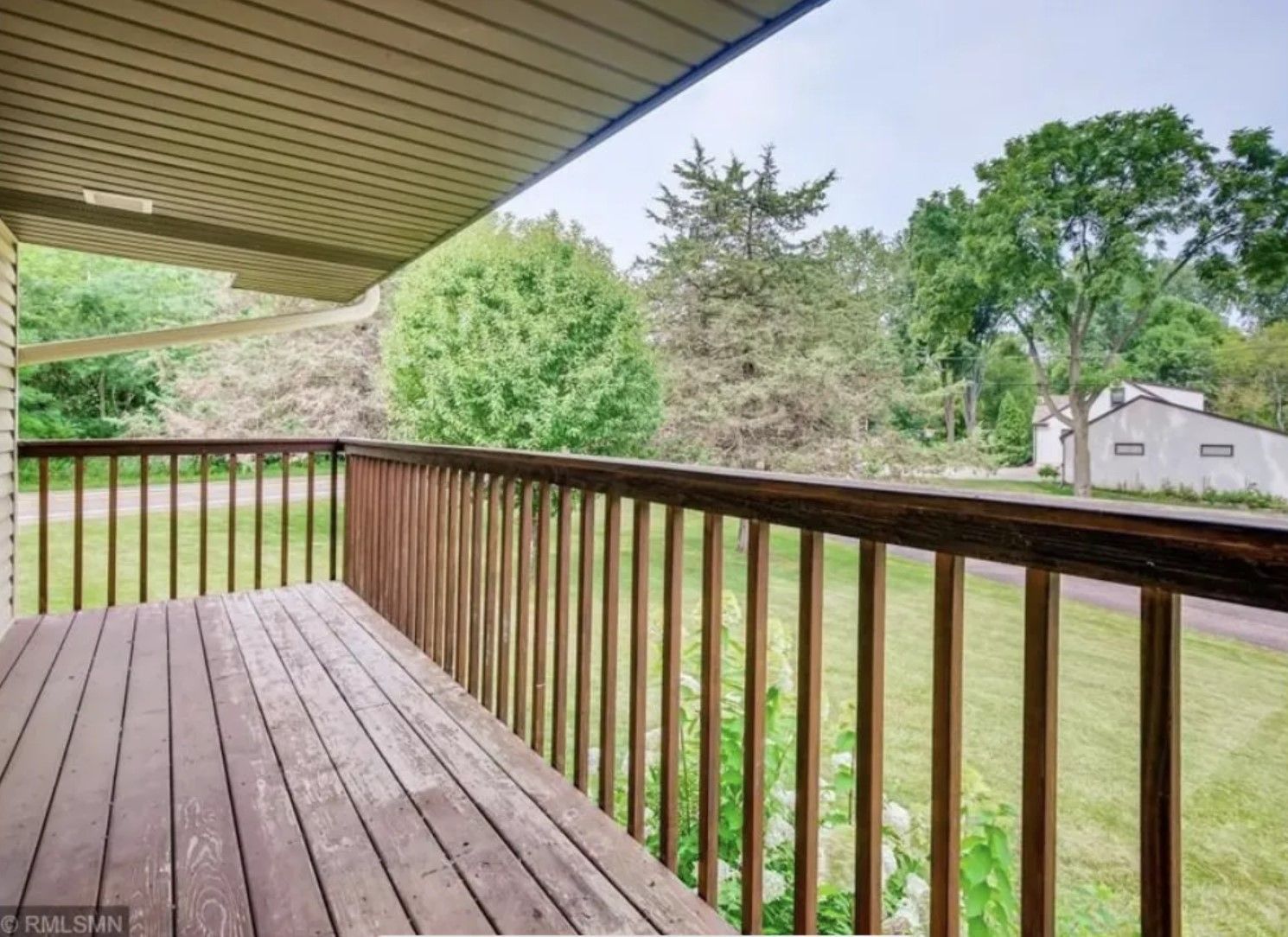 Wooden deck with railing, overlooking a green yard and trees, under a light-colored overhang.