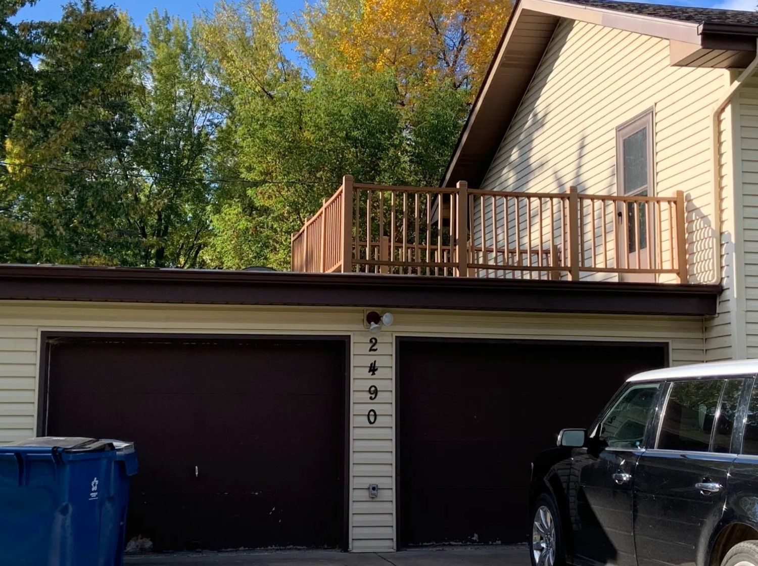 Two-car garage with a brown deck above. The garage doors are brown with the number 2490. A blue trash can sits on the left.