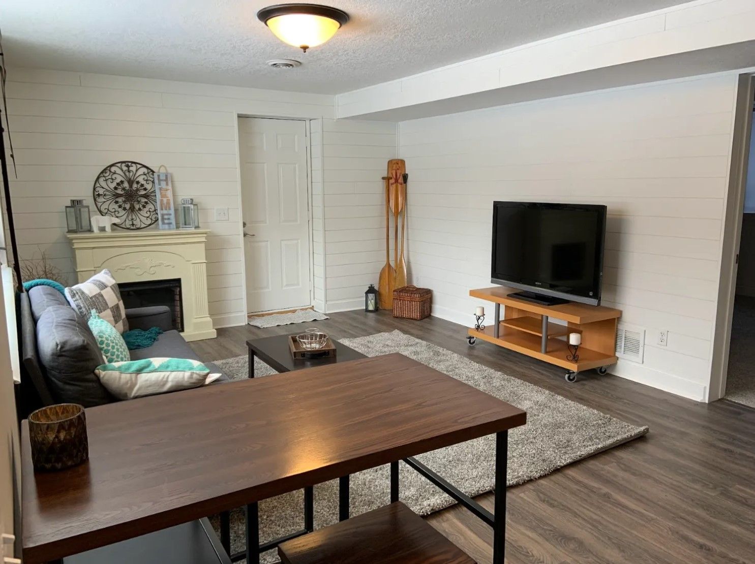 A living room with wood flooring, a tv, sofa, fireplace, and table. Walls are white and wood-paneled.
