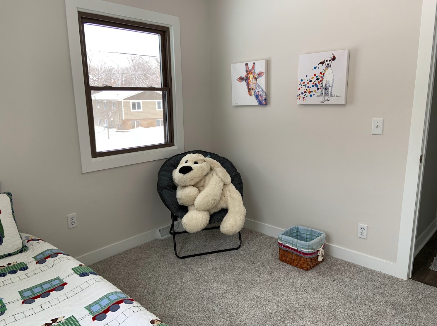 A child's bedroom with a window, chair, plush bear, artwork, and a small woven basket.