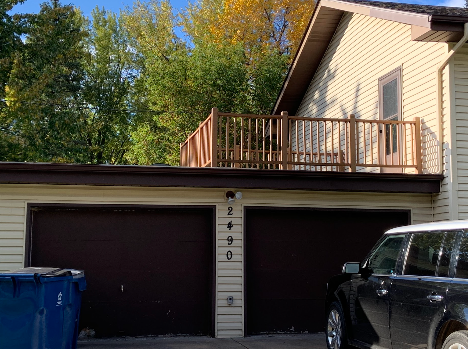 Two-car garage with a deck on the roof, a car parked nearby, and blue bins. Trees and blue sky visible.