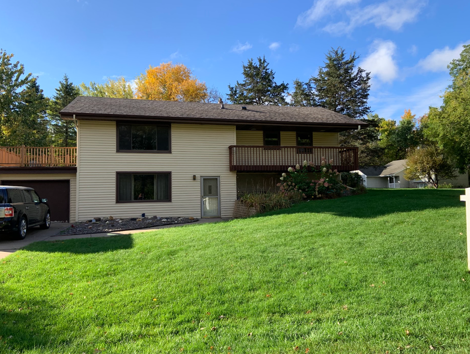 Two-story house with beige siding, brown roof and deck, green lawn, sunny day.