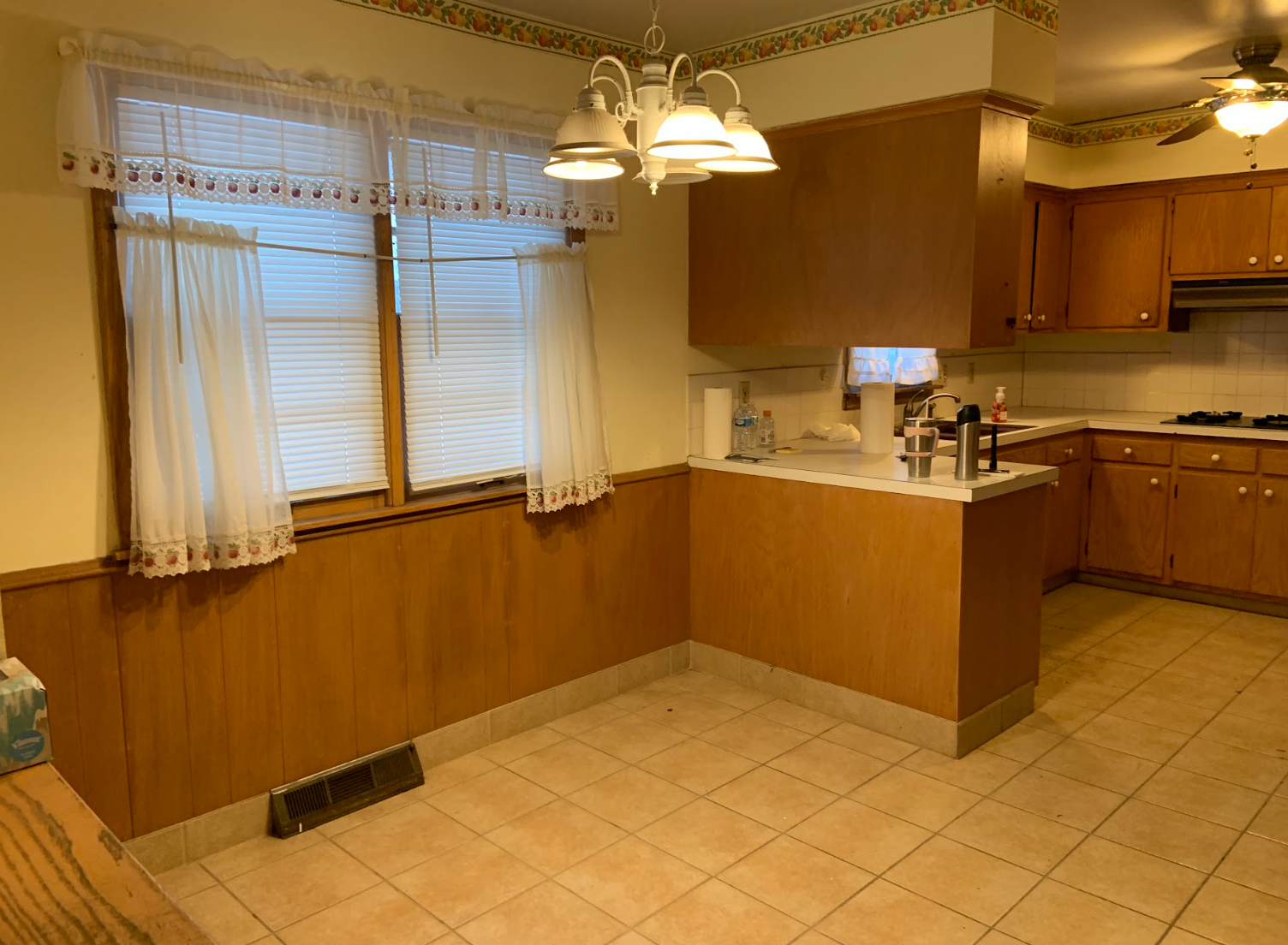 A kitchen with wood paneling, tile floor, and a window with curtains. Cabinets and appliances are visible.