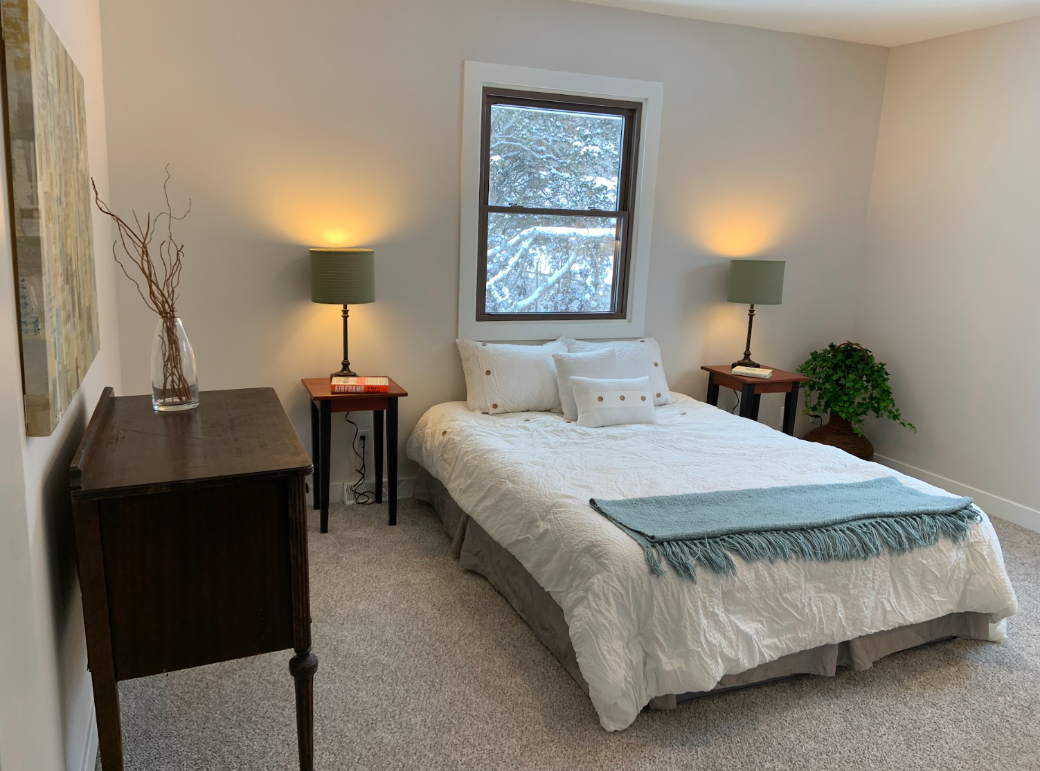 Bedroom with bed, nightstands, lamps, and a window. Light-colored walls and carpet.