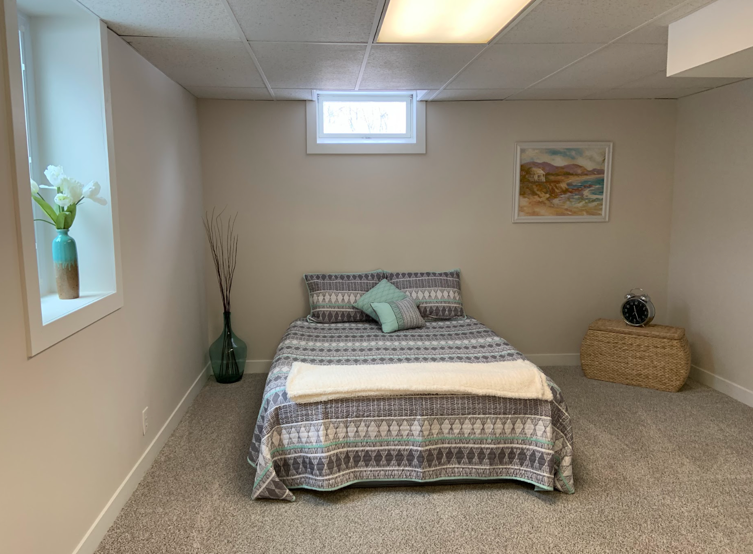 Bedroom with bed, small window, art, woven side table, and beige walls.