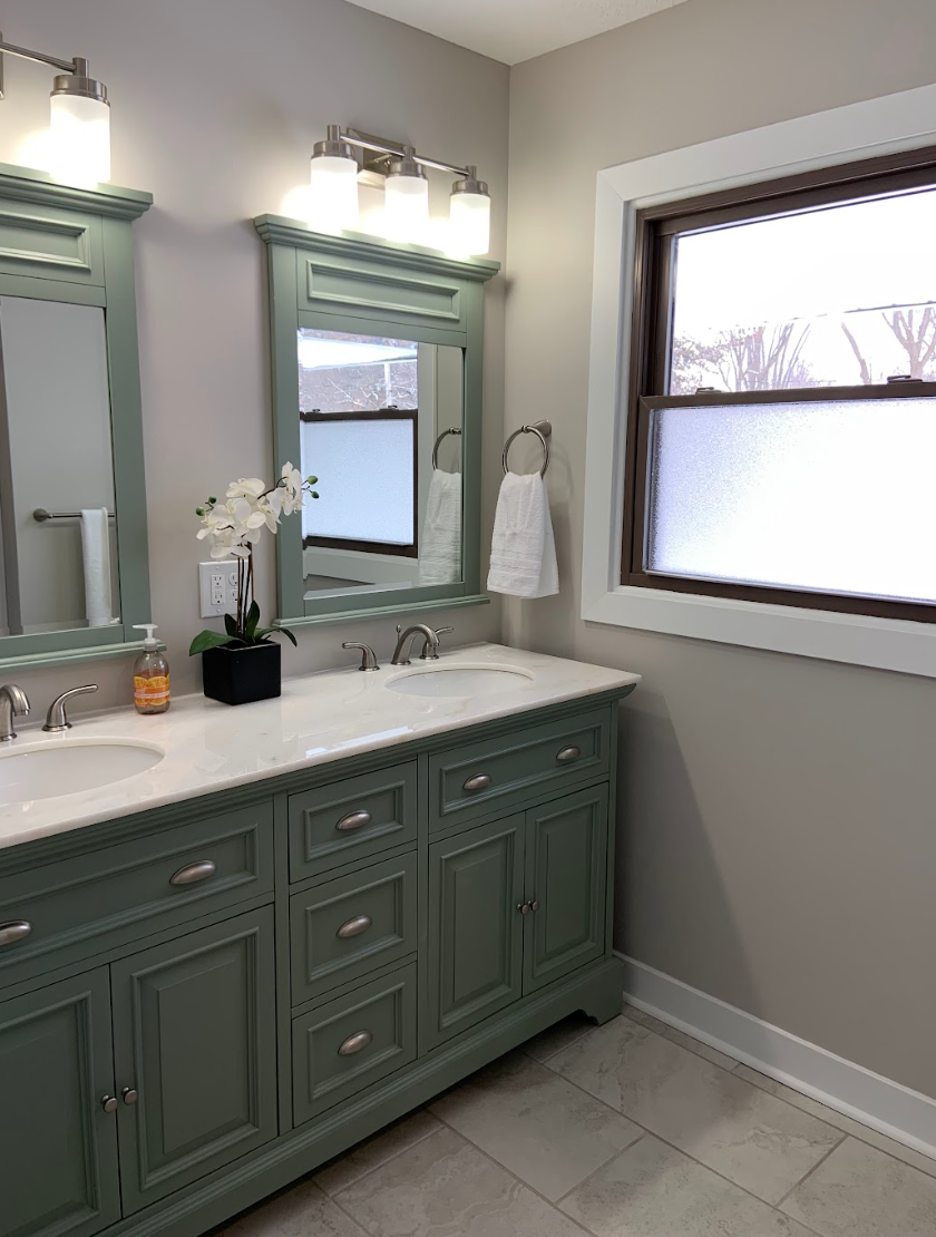 Green double vanity with mirrors in a bathroom, next to a window.