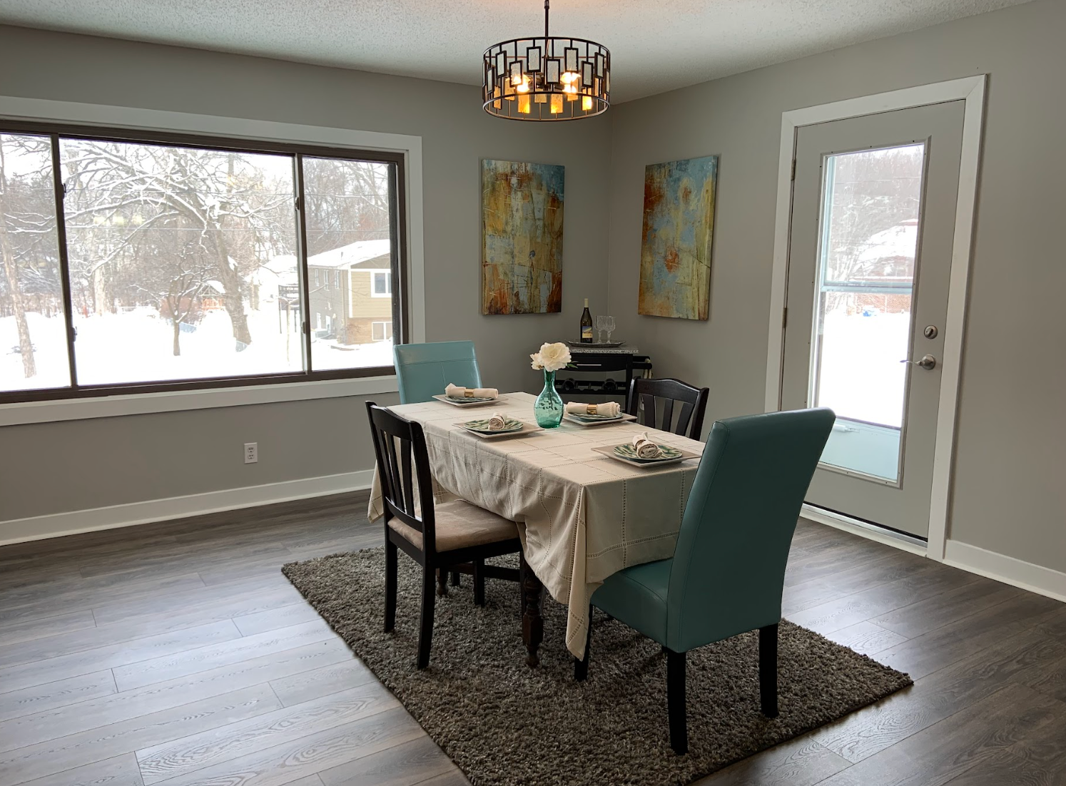 Dining room with table, chairs, art, and a chandelier; large window with snowy outdoor view.