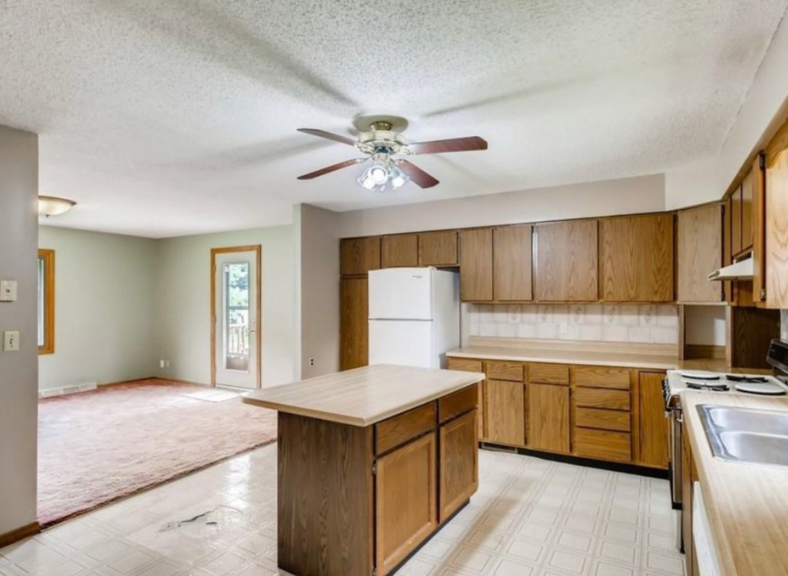 Open-concept kitchen and living area with wood cabinets, island, white refrigerator, and pinkish carpet.