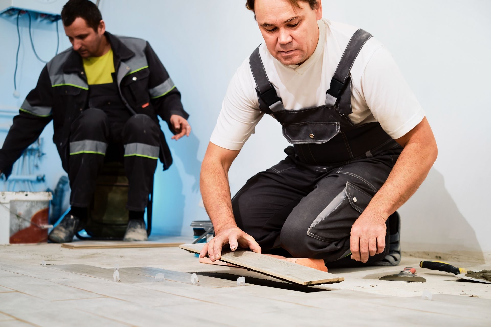 Two workers installing flooring; one kneeling, examining a tile, the other in background.