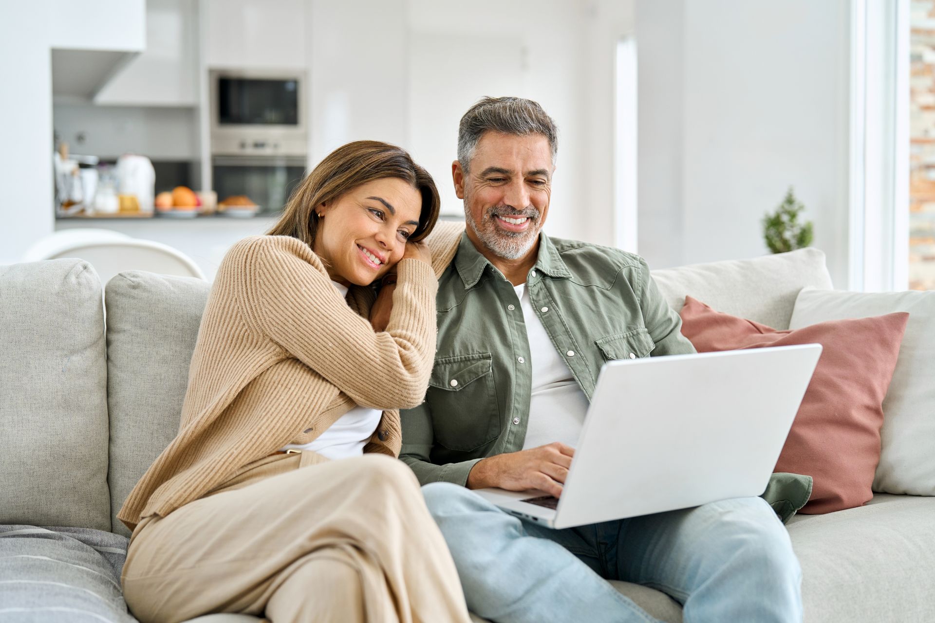 Couple sitting on a couch, looking at a laptop together, smiling. Living room setting.