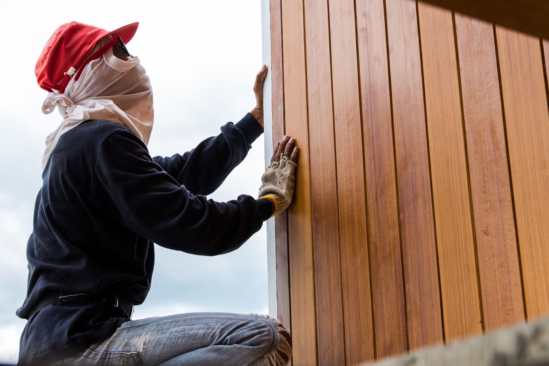 Person installing wooden paneling, wearing a hat, face covering, gloves, against a cloudy sky.