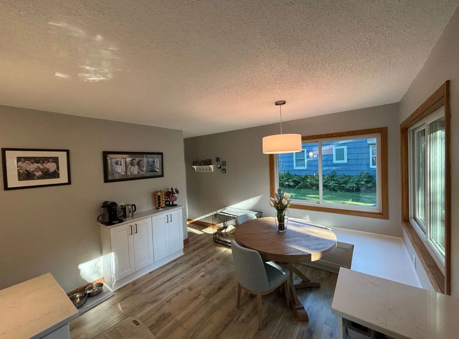Dining room with round table, chairs, and white cabinet. Large window, light-colored walls and flooring.