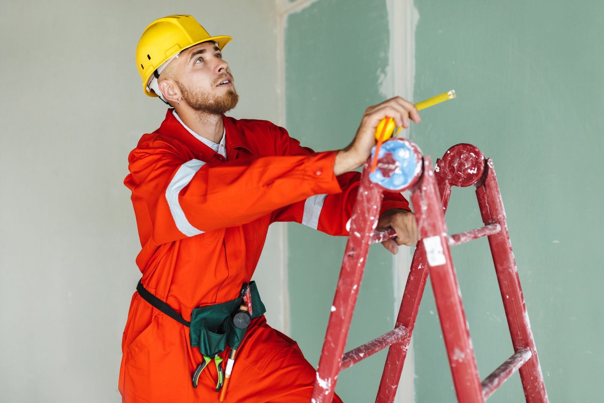 Construction worker on a ladder, measuring a wall with a pencil. He wears orange coveralls and a yellow hard hat.