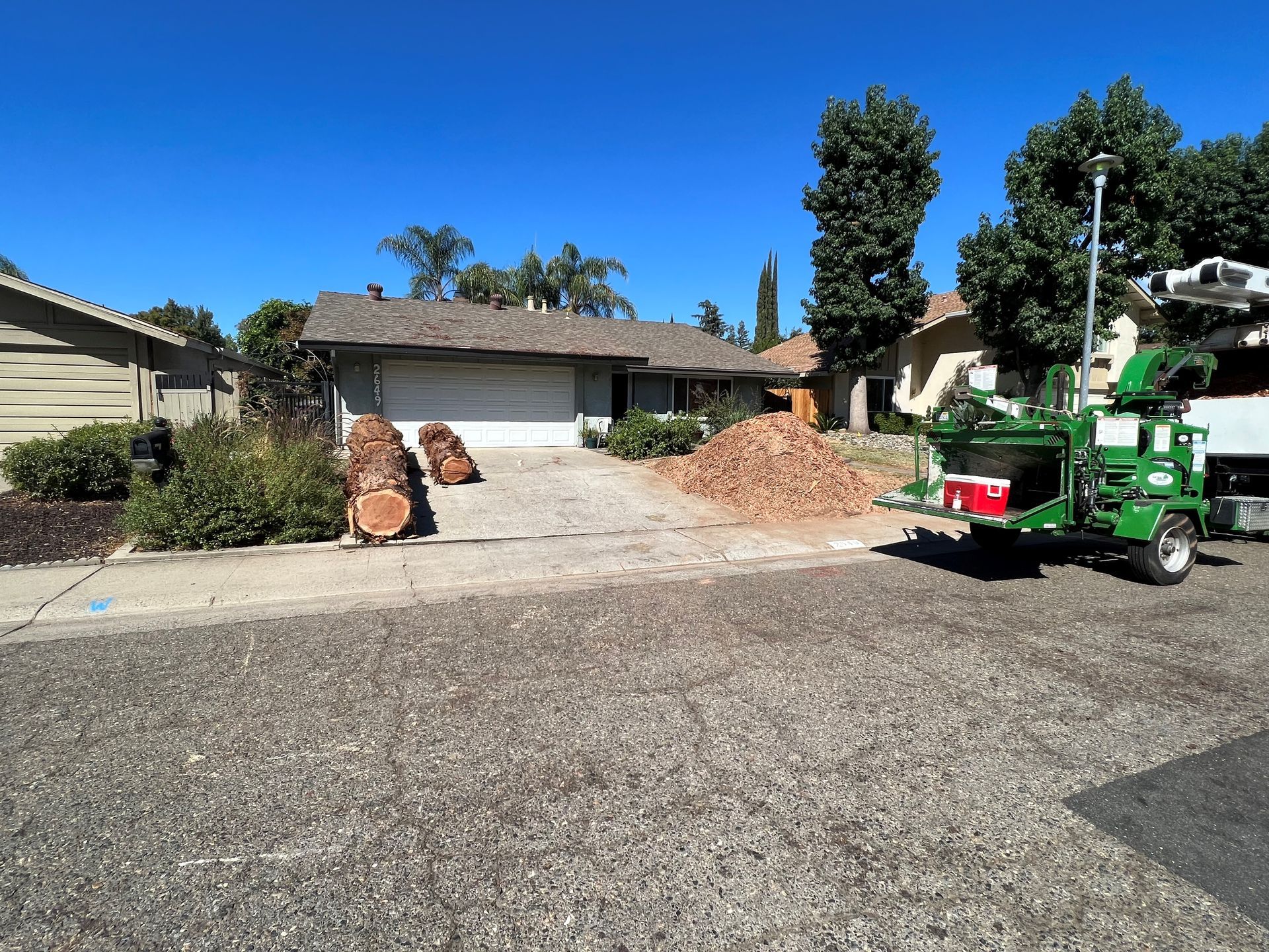 A house with a wood chipper processing logs on the driveway on a sunny day.