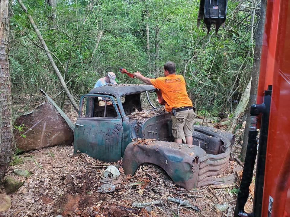 Two people working on a rusted car stuck in a muddy forest clearing, one in an orange shirt.