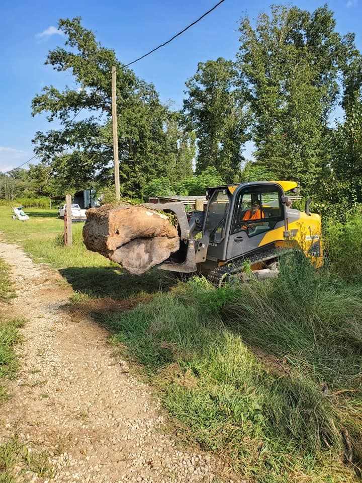 Yellow loader lifting a large tree stump beside a dirt road in a grassy rural area