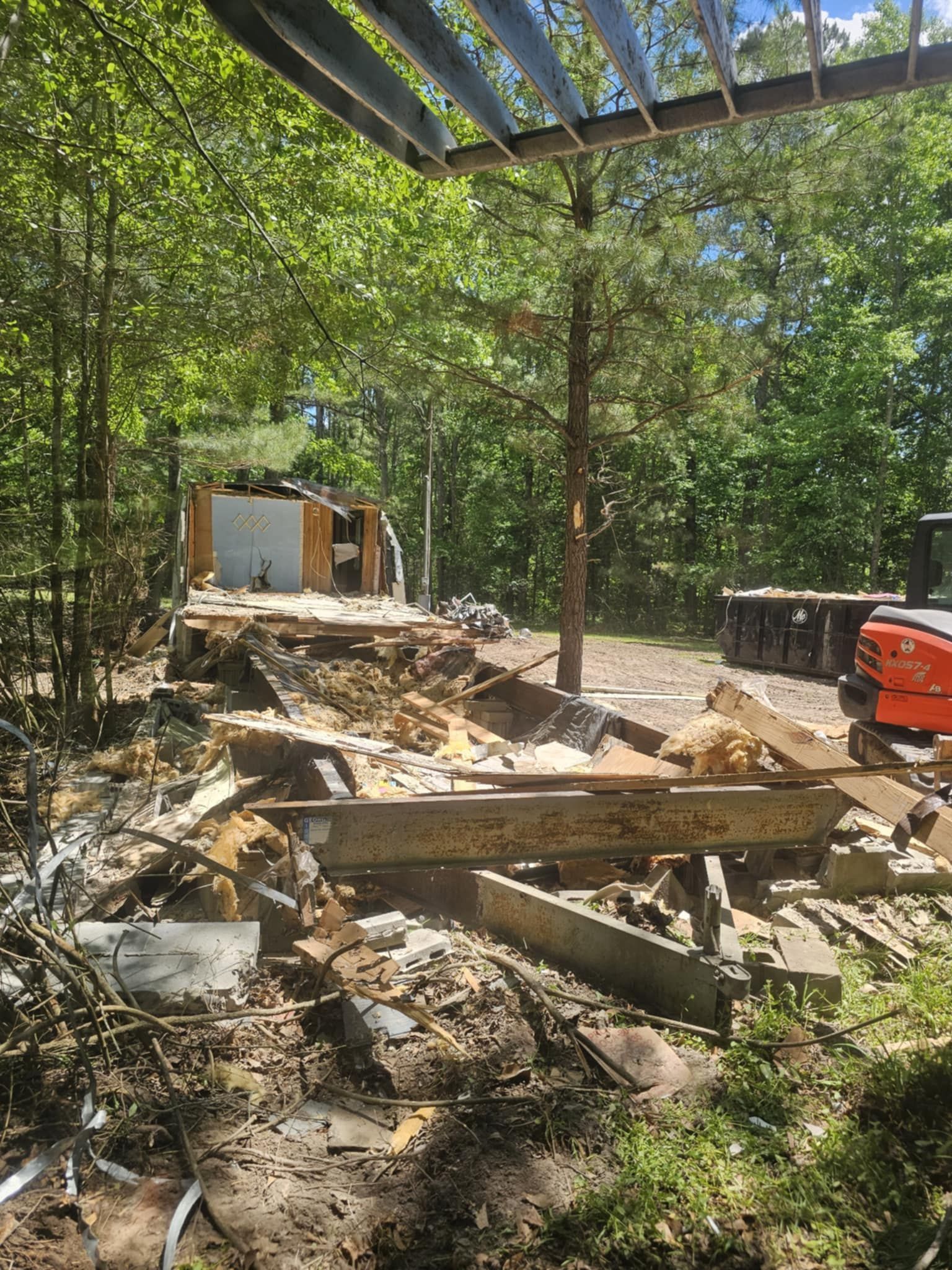 Outdoor construction site with debris, wooden beams, and a red machine under trees near a small shed