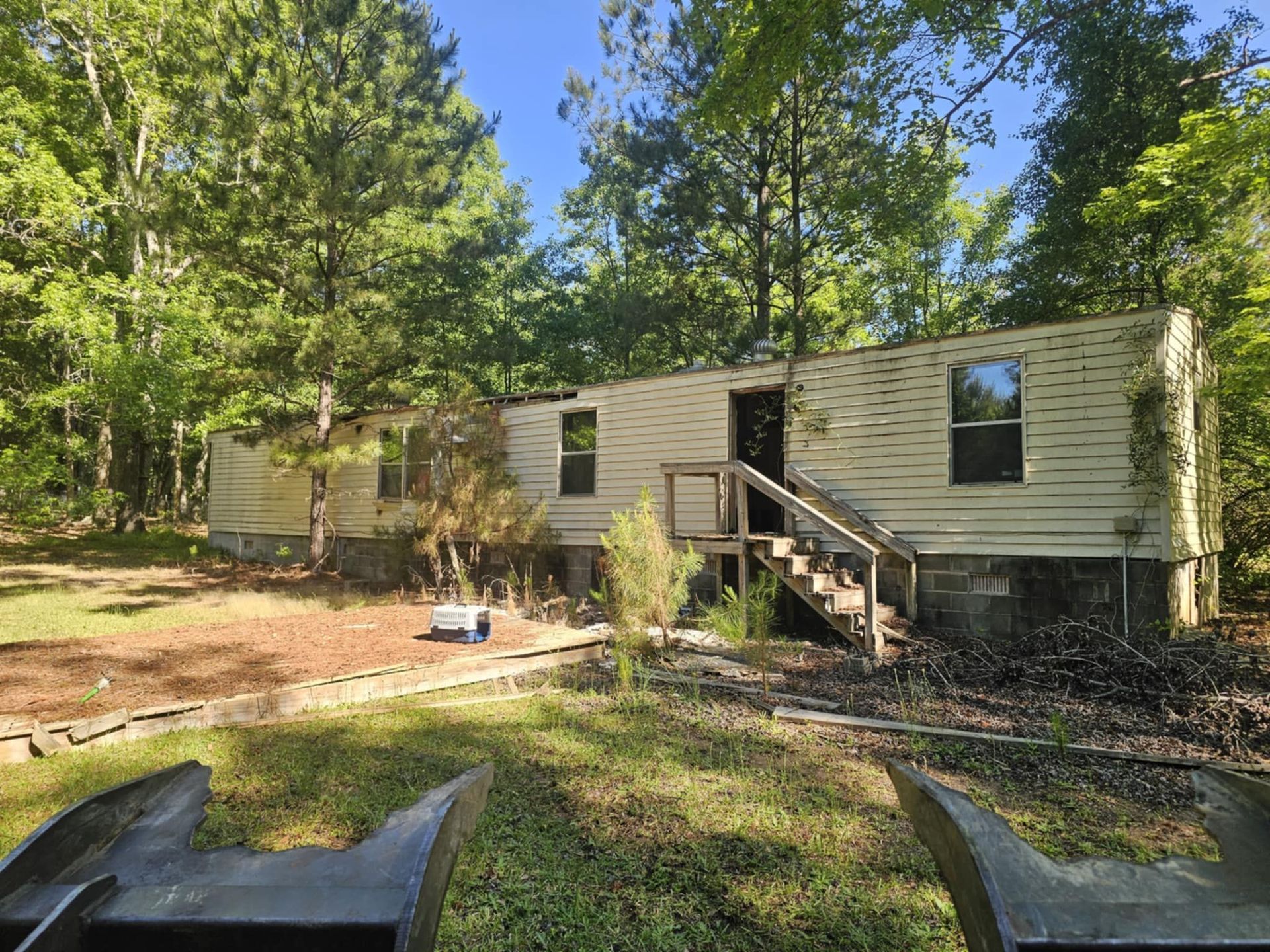 Mobile home in wooded clearing with front steps, windows, and a small yard under tall trees