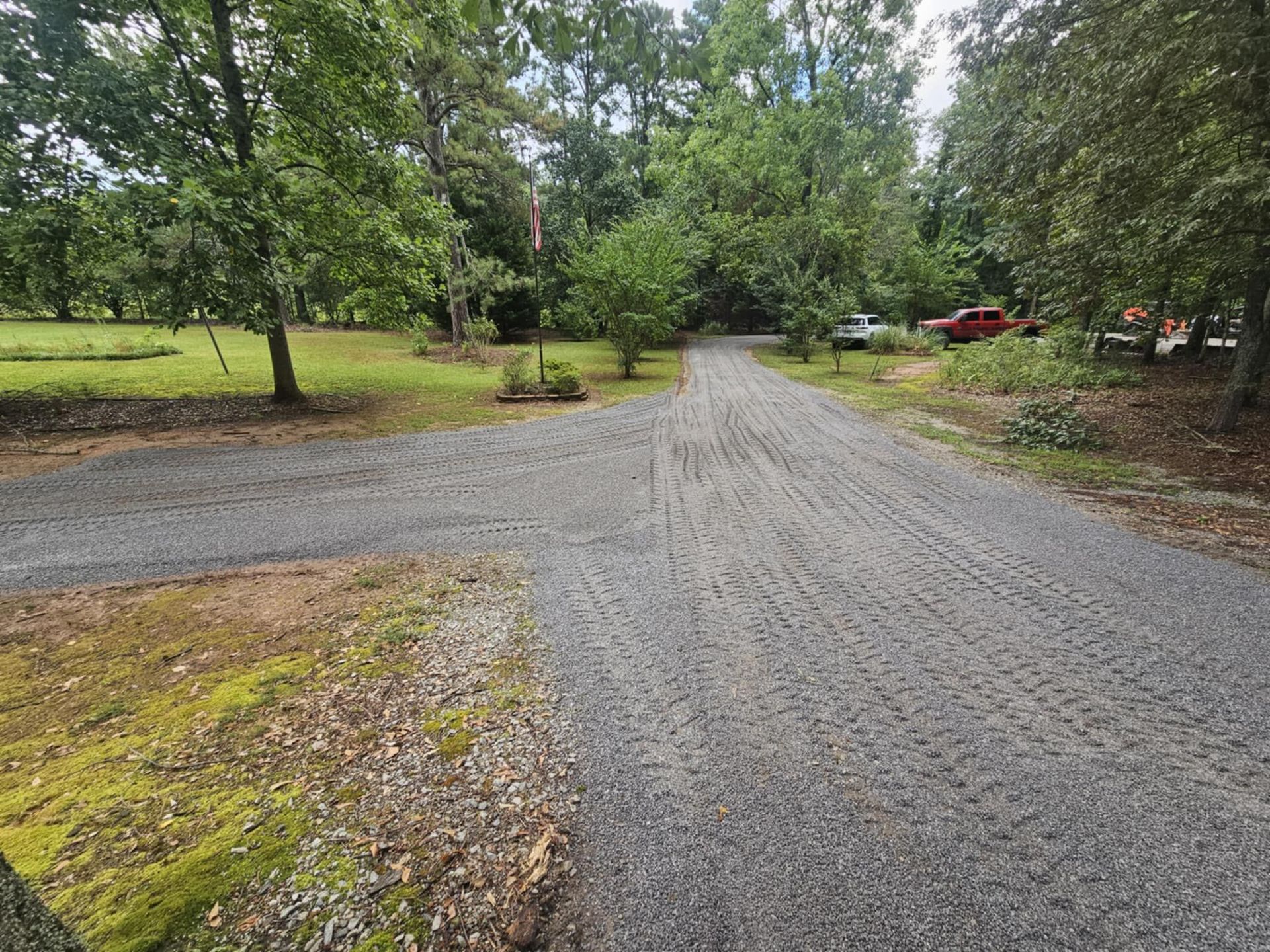 Gravel driveway curving through a wooded yard with green grass and trees.