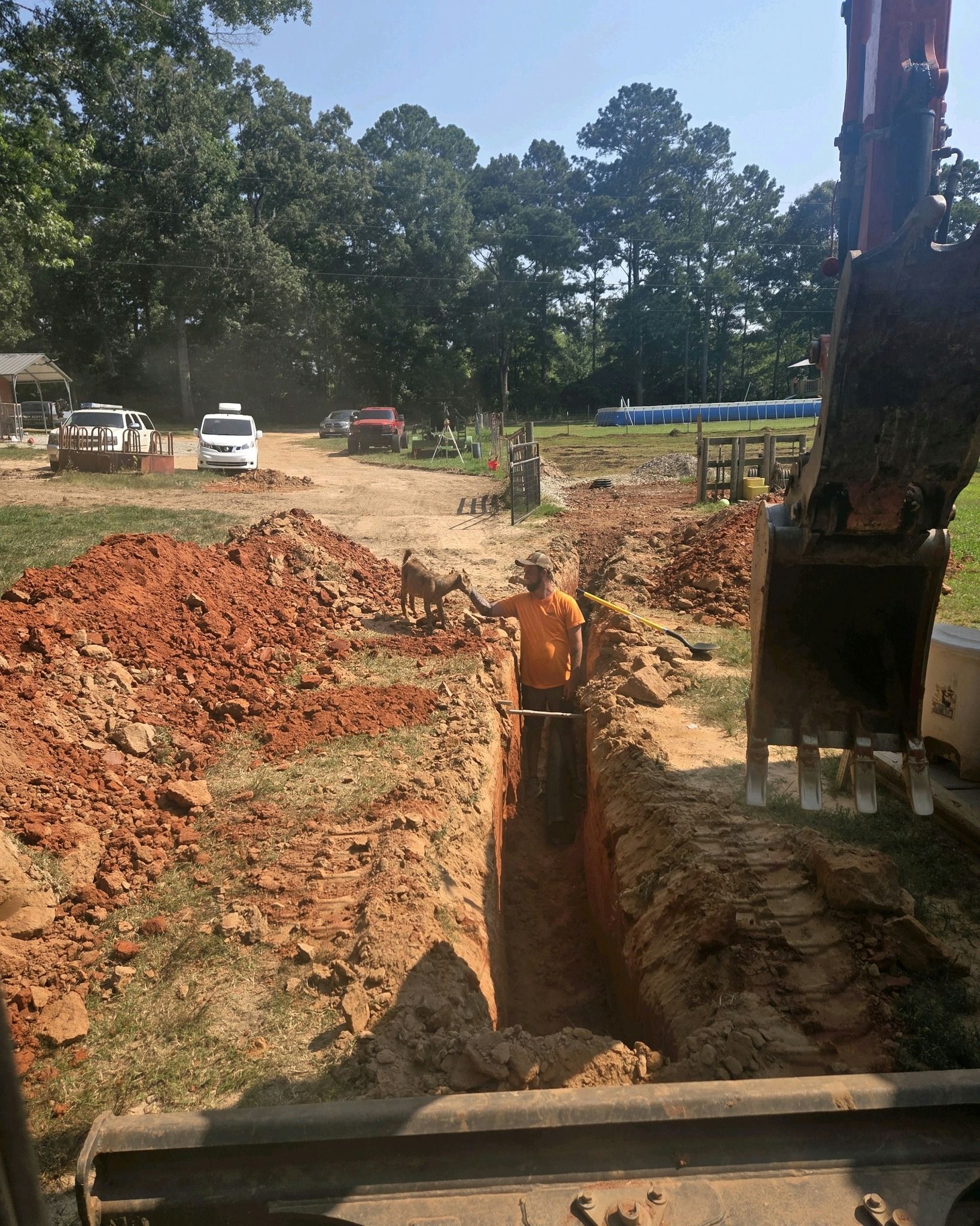 Construction site with excavator digging a trench beside piles of red soil and trees in the background