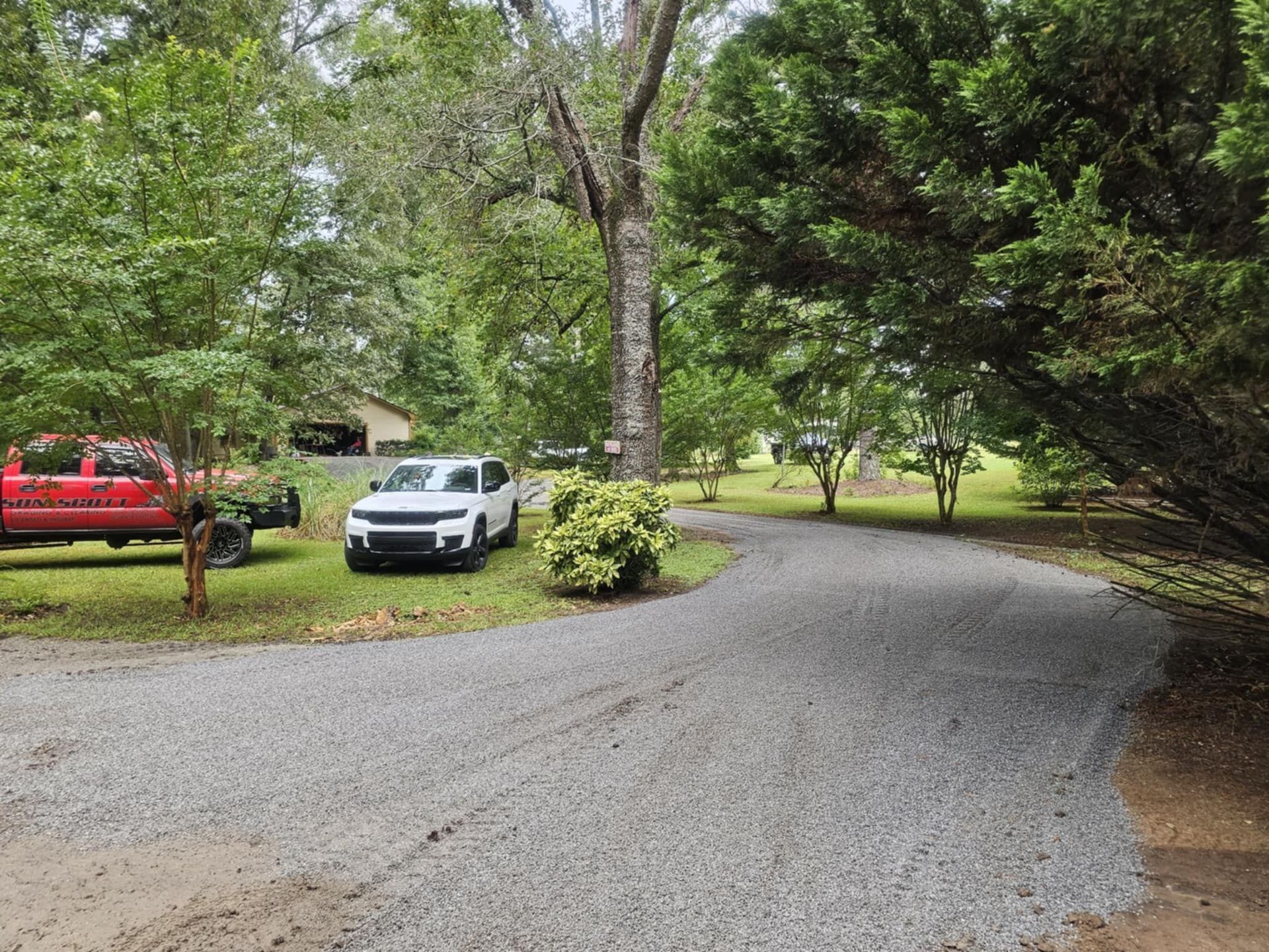 Gravel driveway curving through a wooded yard with a white car and red truck parked nearby