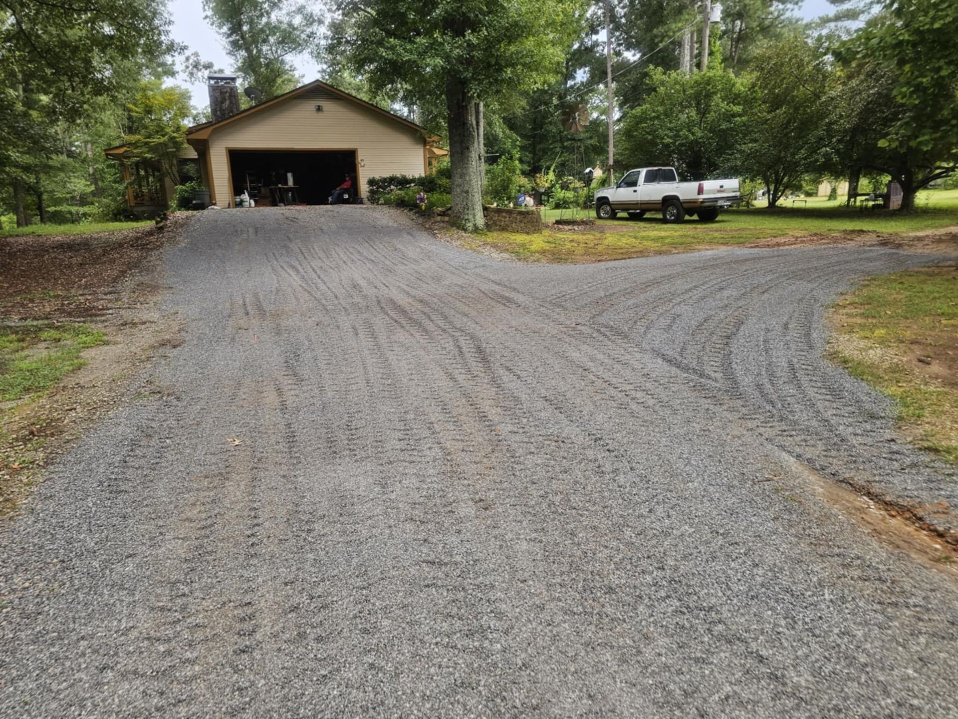 Gravel driveway leading to a garage with a white pickup truck parked beside it, surrounded by trees and grass