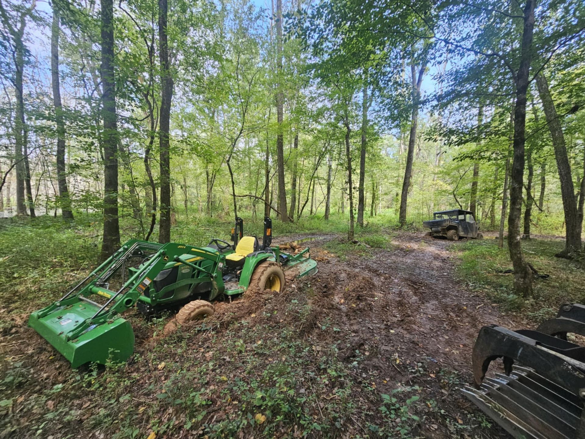 Green tractor with front loader in a wooded dirt clearing, with a black machine at right.