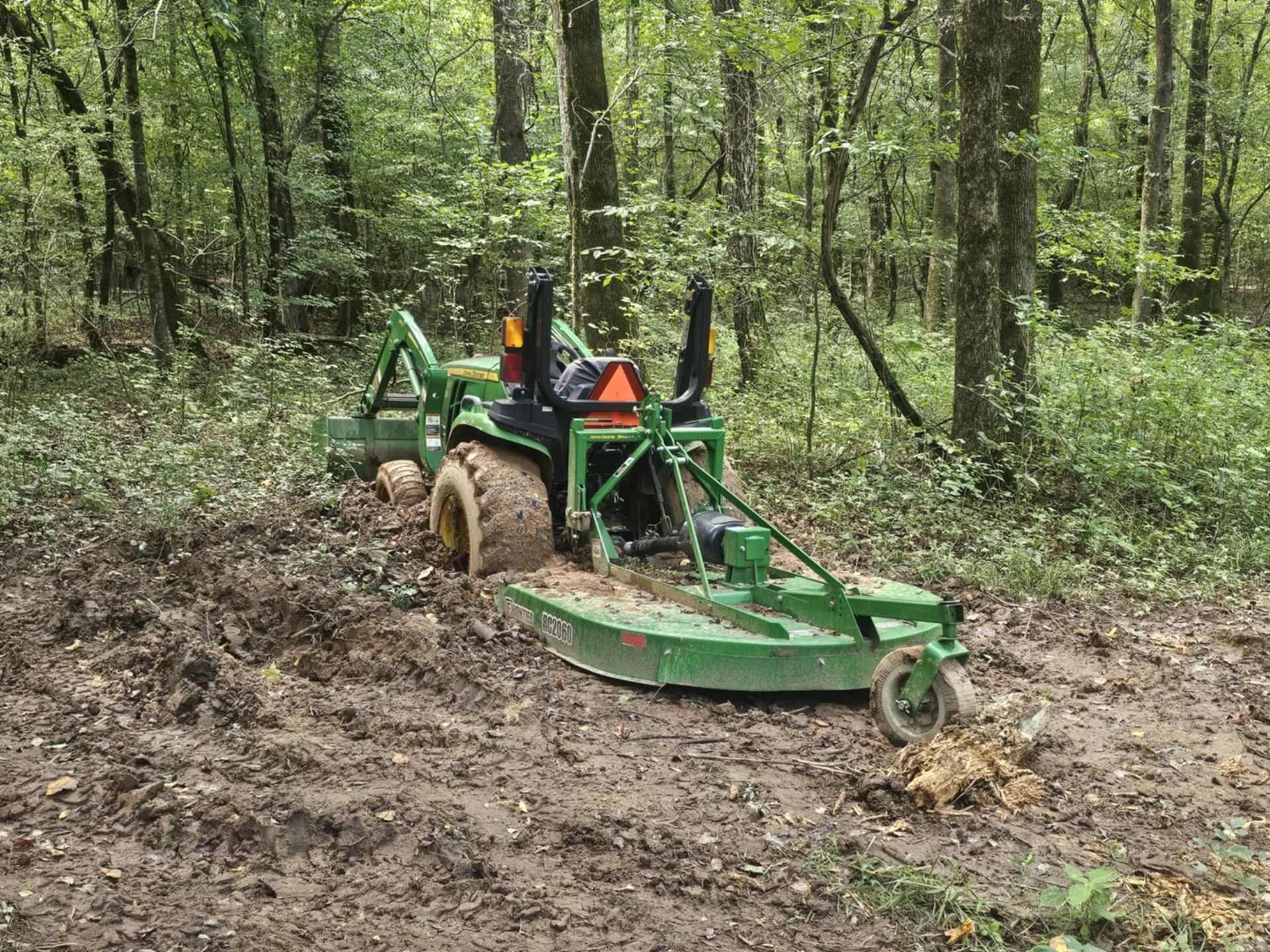 Green tractor mowing muddy forest trail in a wooded area
