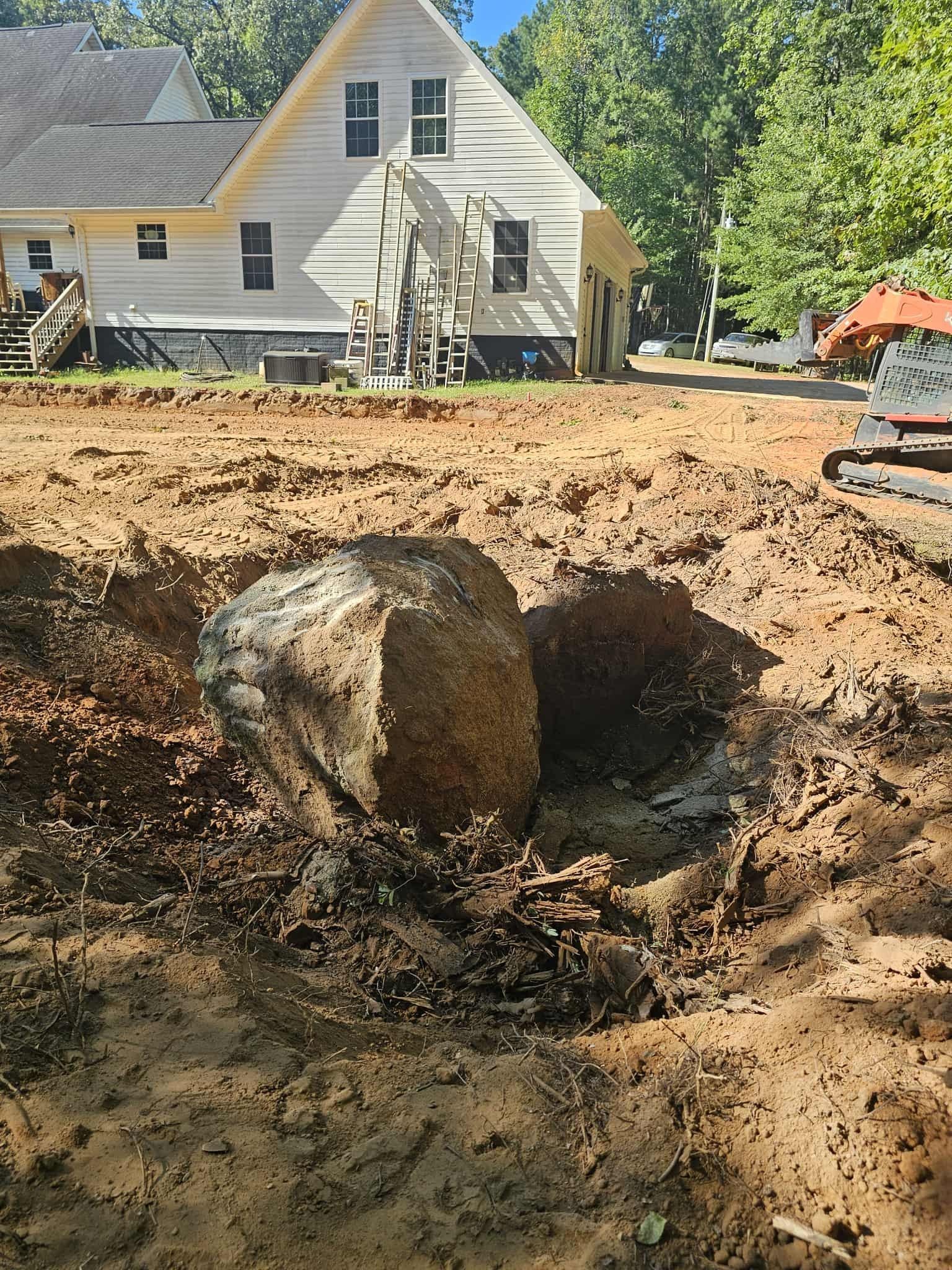 Large boulder in a dug-out hole at a construction site, with a house in the background.