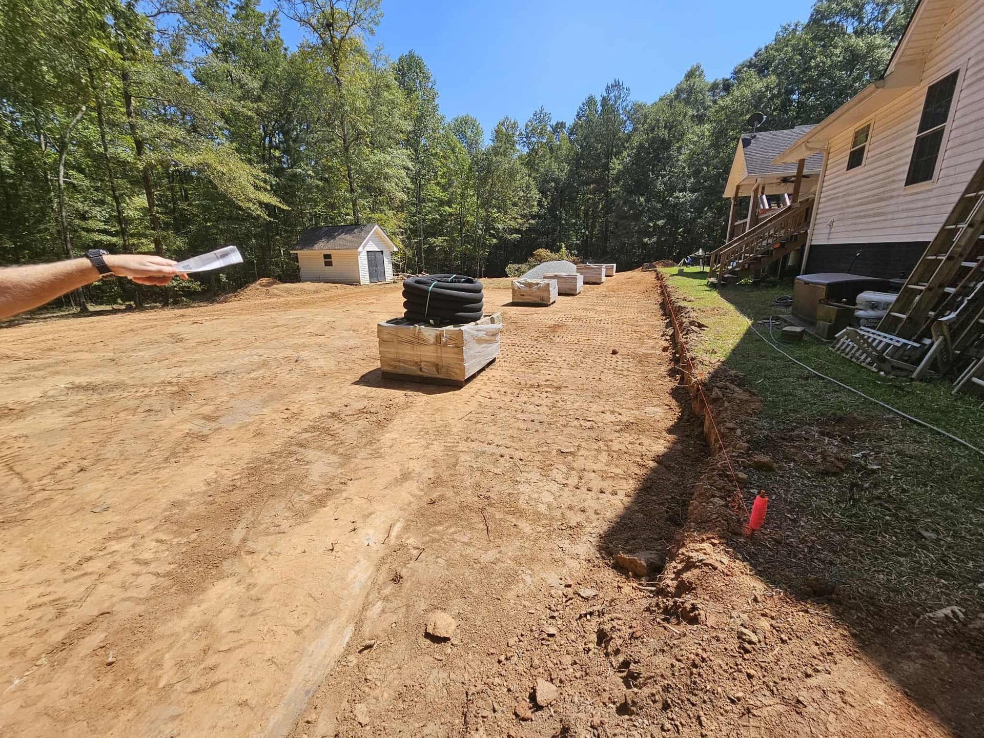 Dirt driveway under construction with stacked bricks, a small house, and trees along the right side.