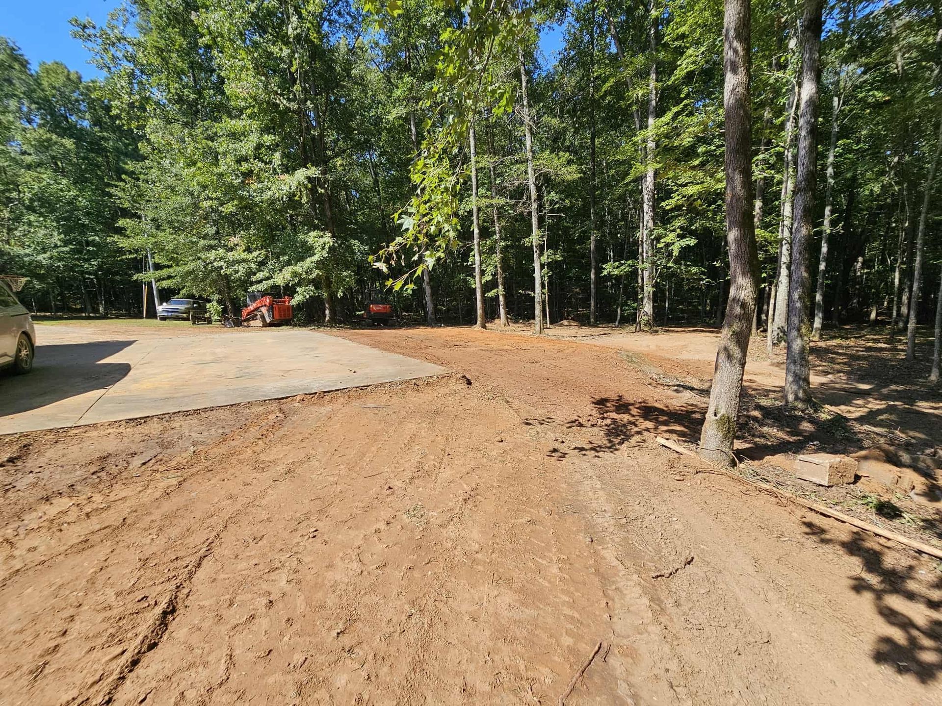 Dirt road under construction beside a paved area and tall trees in a wooded lot.