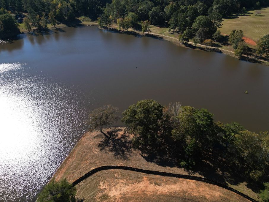 Aerial view of a calm lake with sun glittering on the water, bordered by trees and a dirt path.