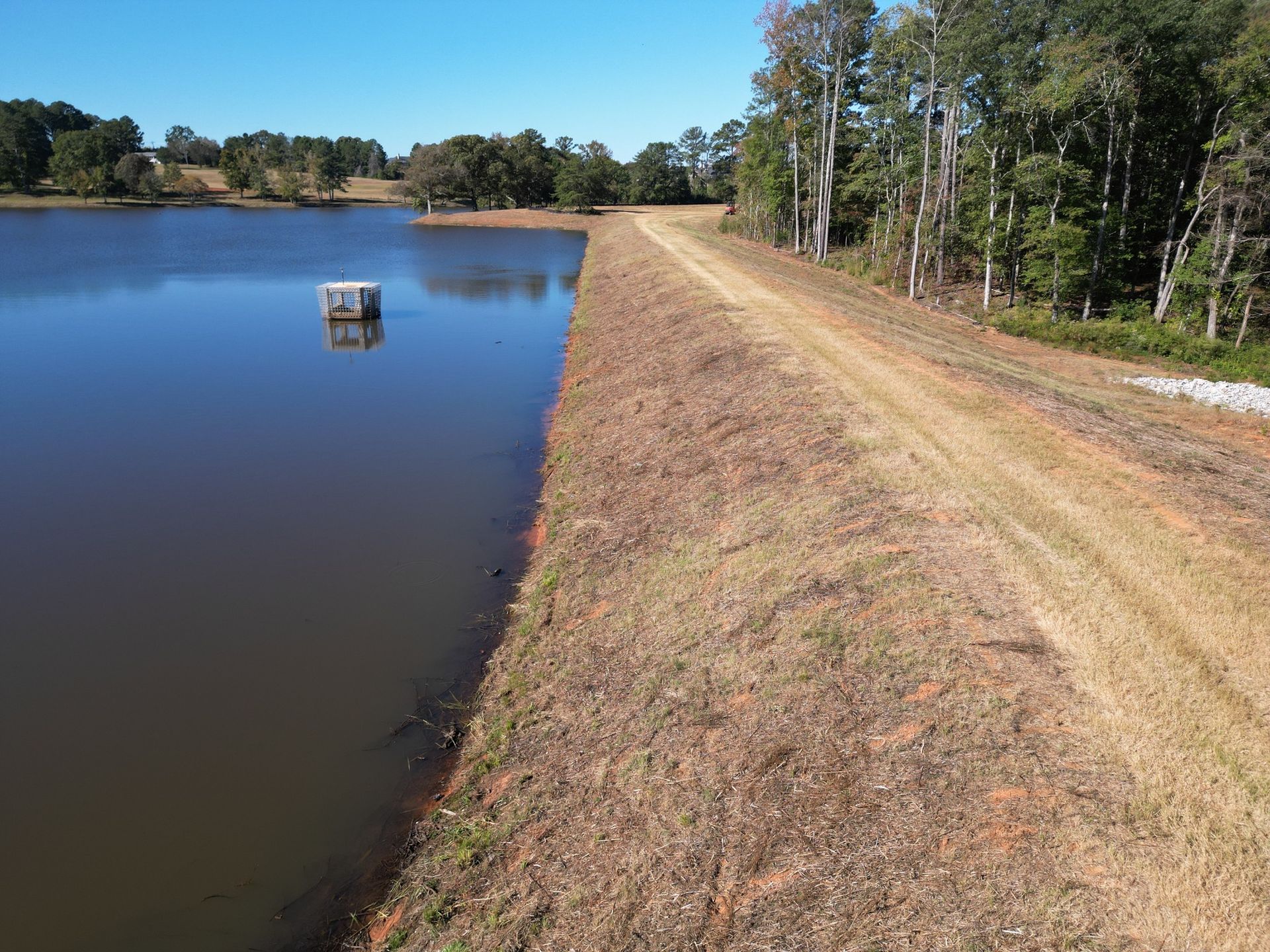 Reservoir beside a dry dirt embankment and tree-lined shore under a clear blue sky