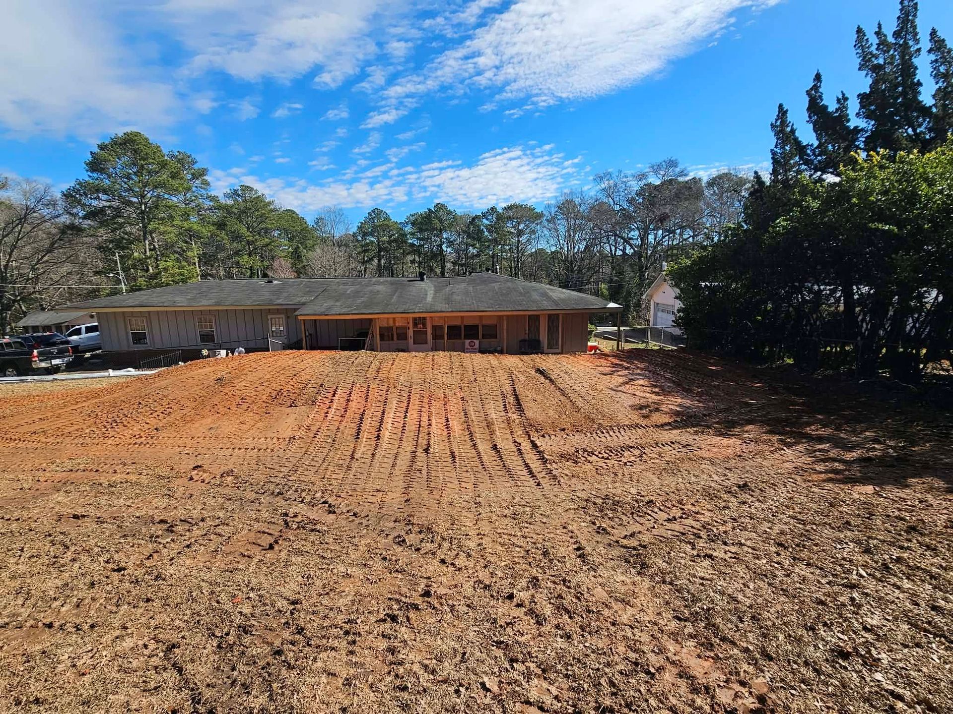 Dirt lot in front of a single-story house under a blue sky, with trees lining the background