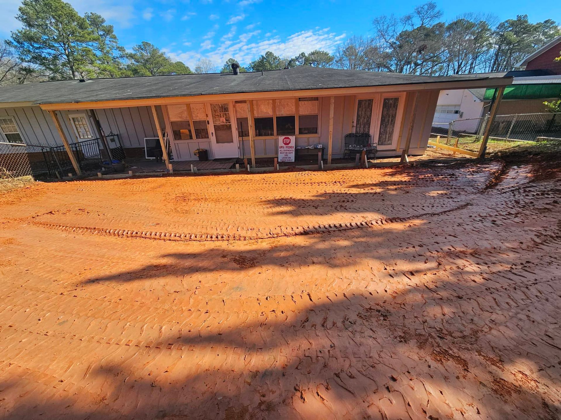 Rural storefront with a covered porch and chairs, set behind a wide red dirt yard under a blue sky