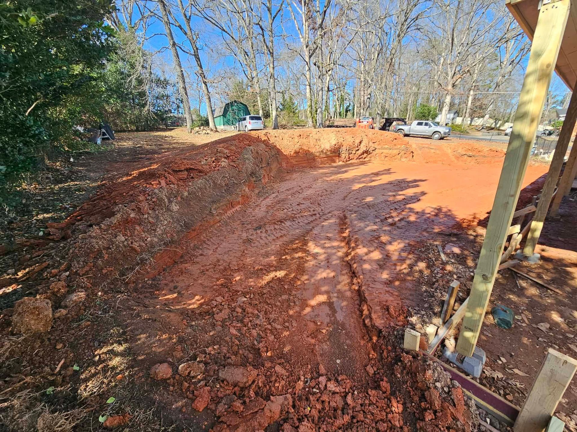 Rutted red dirt road at a construction site beside trees and parked cars
